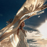 A person stands against a dramatic sky, holding up AYA Sacred Wear's Unique Off-White Boho Poncho with Hand Loomed Tassels that catches the sunlight. The scene exudes a sense of freedom and movement, with the hand loomed tassels adding delicate details as the sun shines brightly in the background.