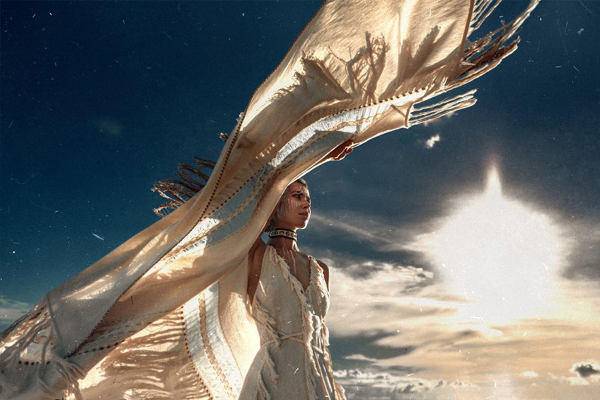 A person stands against a dramatic sky, holding up AYA Sacred Wear's Unique Off-White Boho Poncho with Hand Loomed Tassels that catches the sunlight. The scene exudes a sense of freedom and movement, with the hand loomed tassels adding delicate details as the sun shines brightly in the background.