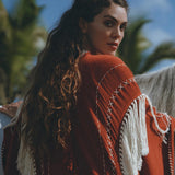 A woman with long, wavy hair stands outside wearing an Unisex Handwoven Cotton Wine Red Poncho from AYA Sacred Wear, featuring a white fringe. She glances back over her shoulder. In the background, palm trees and a blue sky with a few clouds are visible.