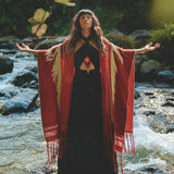 A woman stands on rocks in a flowing stream, arms outstretched. She wears an AYA Sacred Wear Unisex Handwoven Cotton Wine Red Poncho over a black dress. The lush, green setting with sunlight filtering through the trees creates a serene, nature-focused scene.