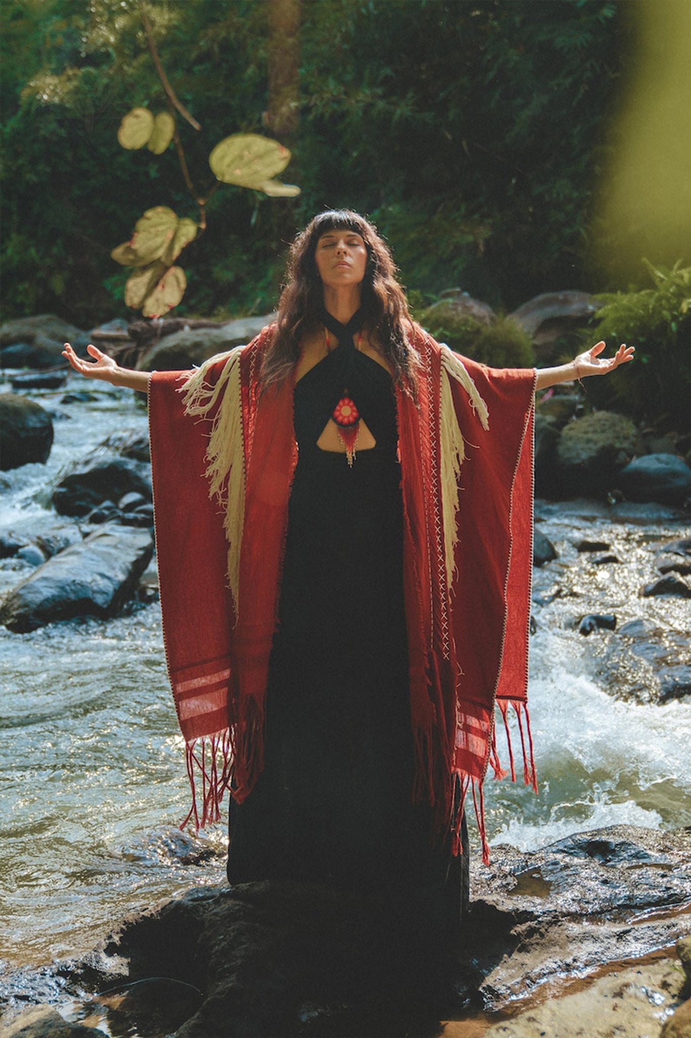 A woman stands on rocks in a flowing stream, arms outstretched. She wears an AYA Sacred Wear Unisex Handwoven Cotton Wine Red Poncho over a black dress. The lush, green setting with sunlight filtering through the trees creates a serene, nature-focused scene.