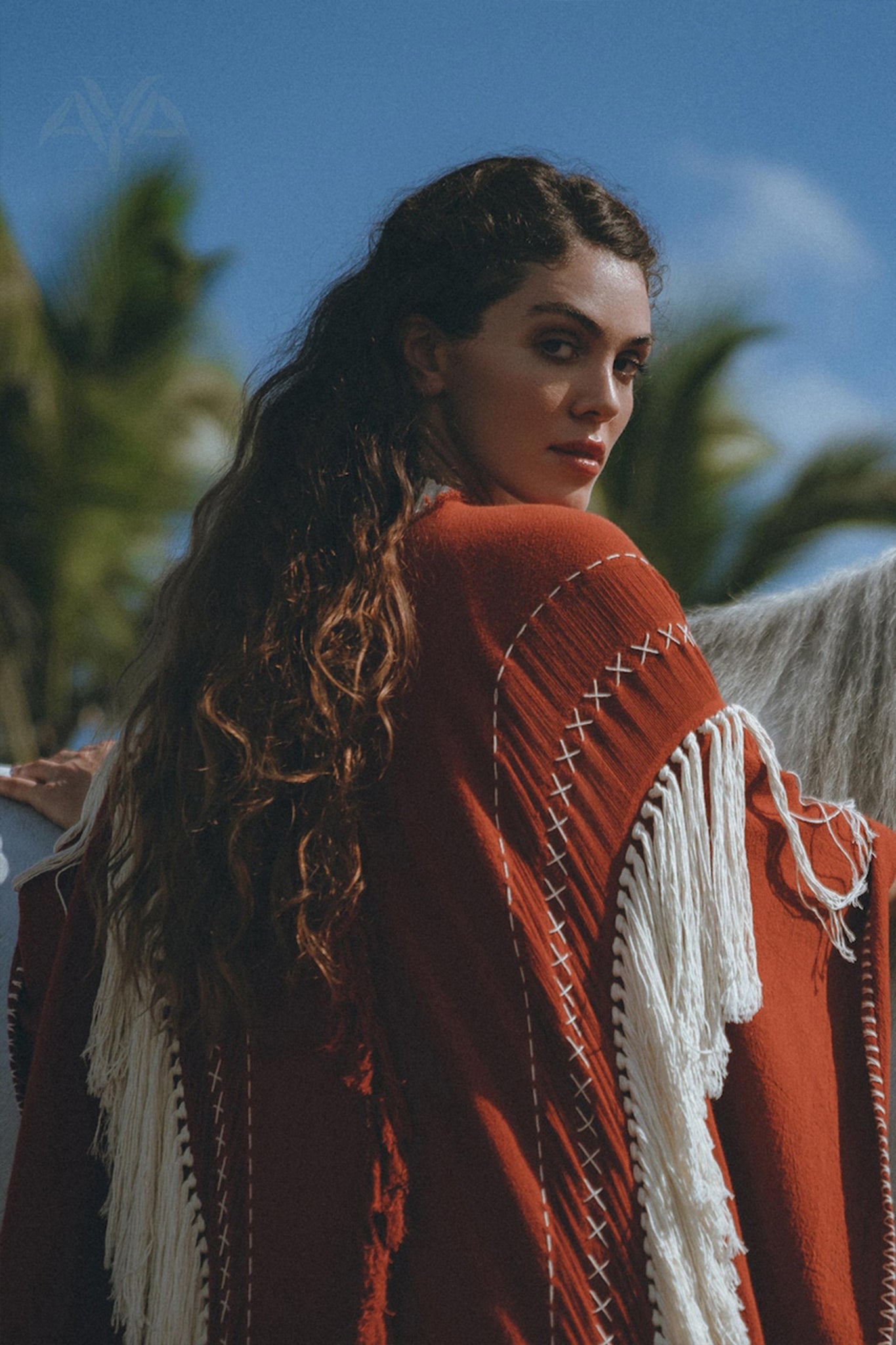 A woman with long, wavy hair stands outside wearing an Unisex Handwoven Cotton Wine Red Poncho from AYA Sacred Wear, featuring a white fringe. She glances back over her shoulder. In the background, palm trees and a blue sky with a few clouds are visible.