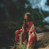 A woman wearing the Unisex Handwoven Cotton Wine Red Poncho from AYA Sacred Wear sits on a rock amidst lush greenery. She looks upwards as sunlight filters through the trees, creating a peaceful and natural atmosphere around her.