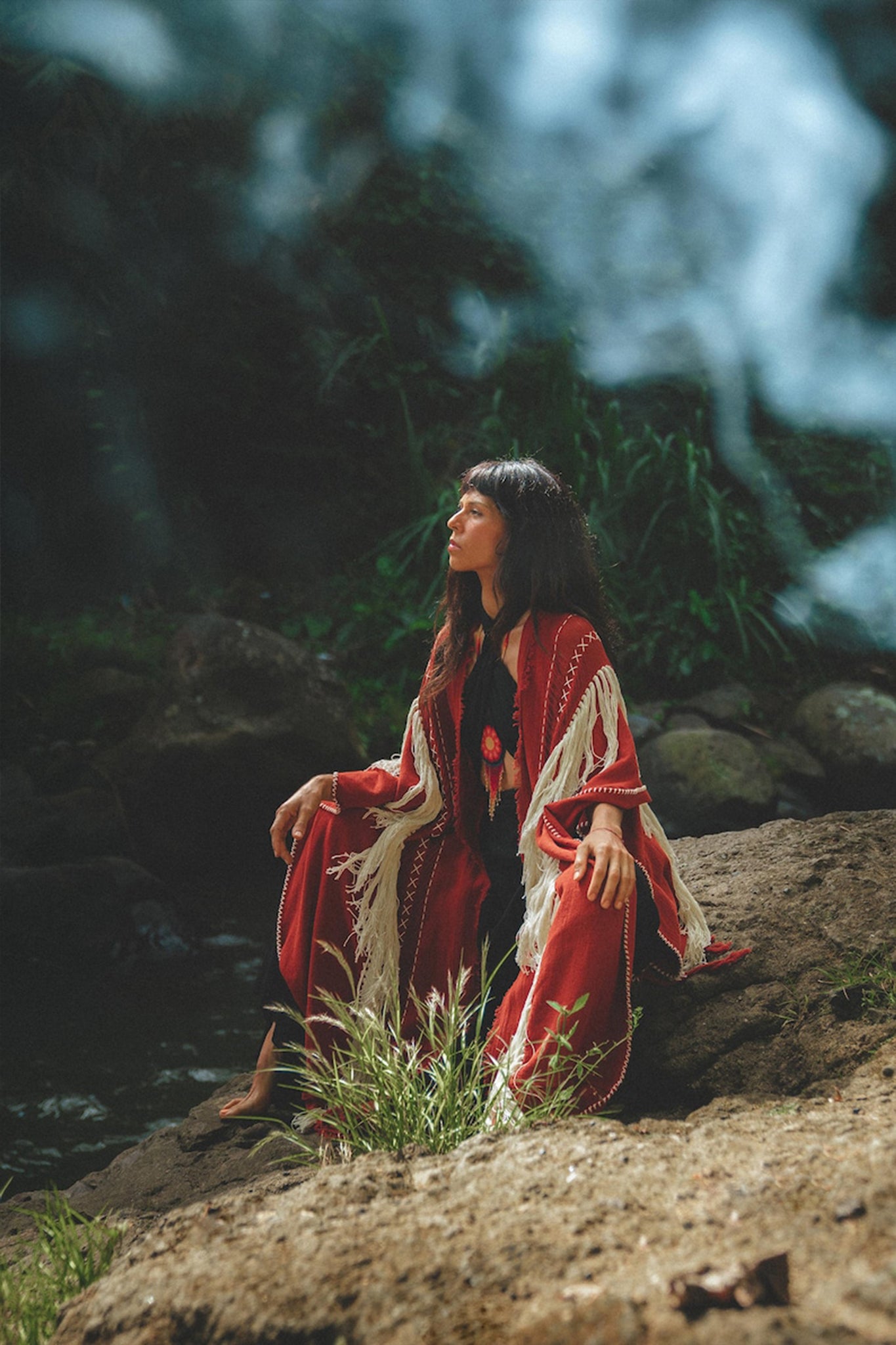 A woman wearing the Unisex Handwoven Cotton Wine Red Poncho from AYA Sacred Wear sits on a rock amidst lush greenery. She looks upwards as sunlight filters through the trees, creating a peaceful and natural atmosphere around her.