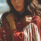 A person with long dark hair is standing outdoors, wrapped in the Unisex Handwoven Cotton Wine Red Poncho by AYA Sacred Wear. They have a thoughtful expression and are looking directly at the camera. The poncho, crafted from organic cotton, showcases intricate stitching and tassel details.