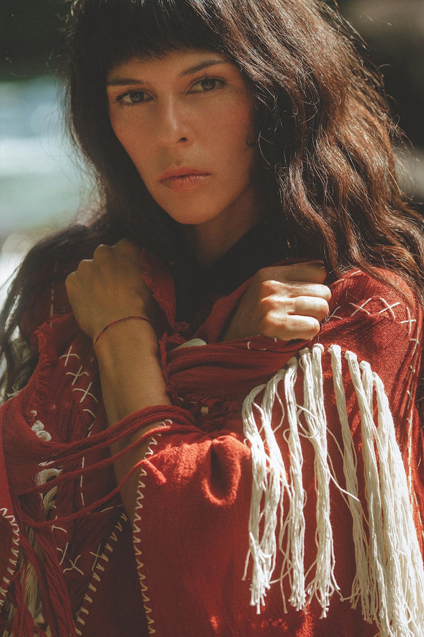 A person with long dark hair is standing outdoors, wrapped in the Unisex Handwoven Cotton Wine Red Poncho by AYA Sacred Wear. They have a thoughtful expression and are looking directly at the camera. The poncho, crafted from organic cotton, showcases intricate stitching and tassel details.