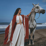 A woman wearing an AYA Sacred Wear organic cotton dress and a Unisex Handwoven Cotton Wine Red Poncho stands elegantly on a beach beside a white horse, with the clear blue sky and ocean waves in the background.