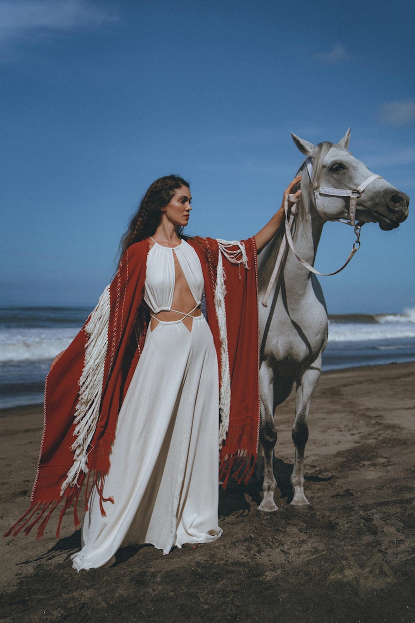 A woman wearing an AYA Sacred Wear organic cotton dress and a Unisex Handwoven Cotton Wine Red Poncho stands elegantly on a beach beside a white horse, with the clear blue sky and ocean waves in the background.