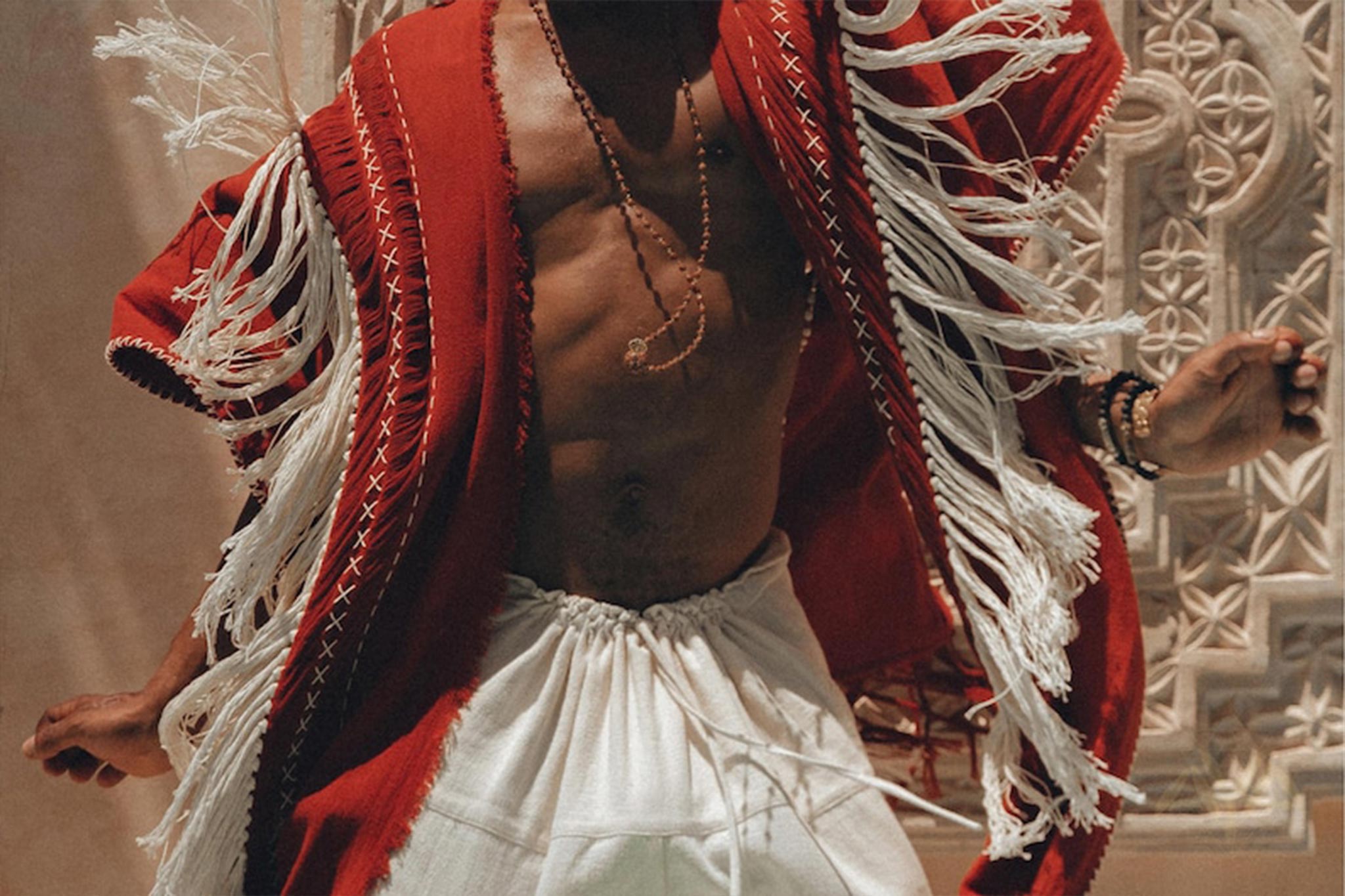 A person dances energetically, draped in an Unisex Handwoven Cotton Wine Red Poncho from AYA Sacred Wear, paired with white pants. The poncho sways dynamically, capturing motion and joy against a backdrop of intricate decorative patterns.