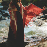 A woman stands gracefully by a flowing stream, enveloped in an AYA Sacred Wear Unisex Handwoven Cotton Wine Red Poncho over her long black dress. She gazes downward, surrounded by lush greenery in a serene, natural setting.