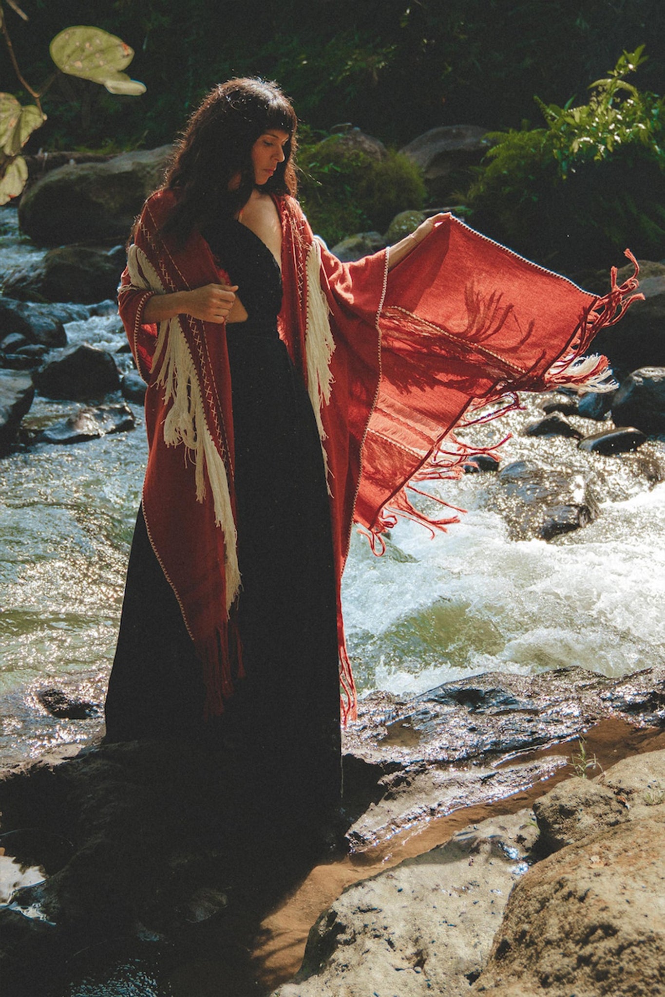 A woman stands gracefully by a flowing stream, enveloped in an AYA Sacred Wear Unisex Handwoven Cotton Wine Red Poncho over her long black dress. She gazes downward, surrounded by lush greenery in a serene, natural setting.