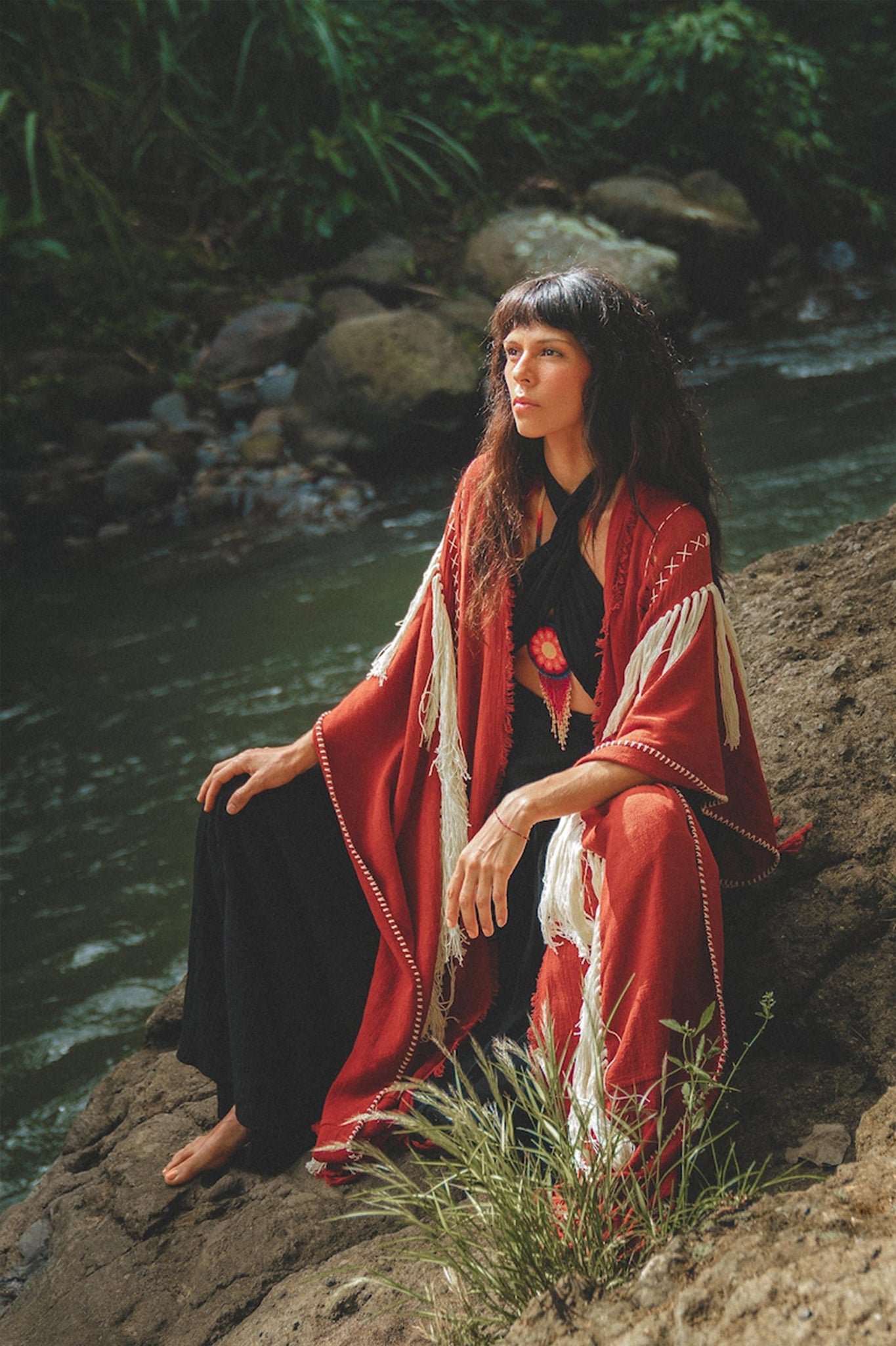 A woman in a wine red handwoven cotton poncho from AYA Sacred Wear sits on a rock by the river, surrounded by lush greenery. She gazes thoughtfully into the distance, her long hair falling over her shoulders. The scene is serene and nature-filled.