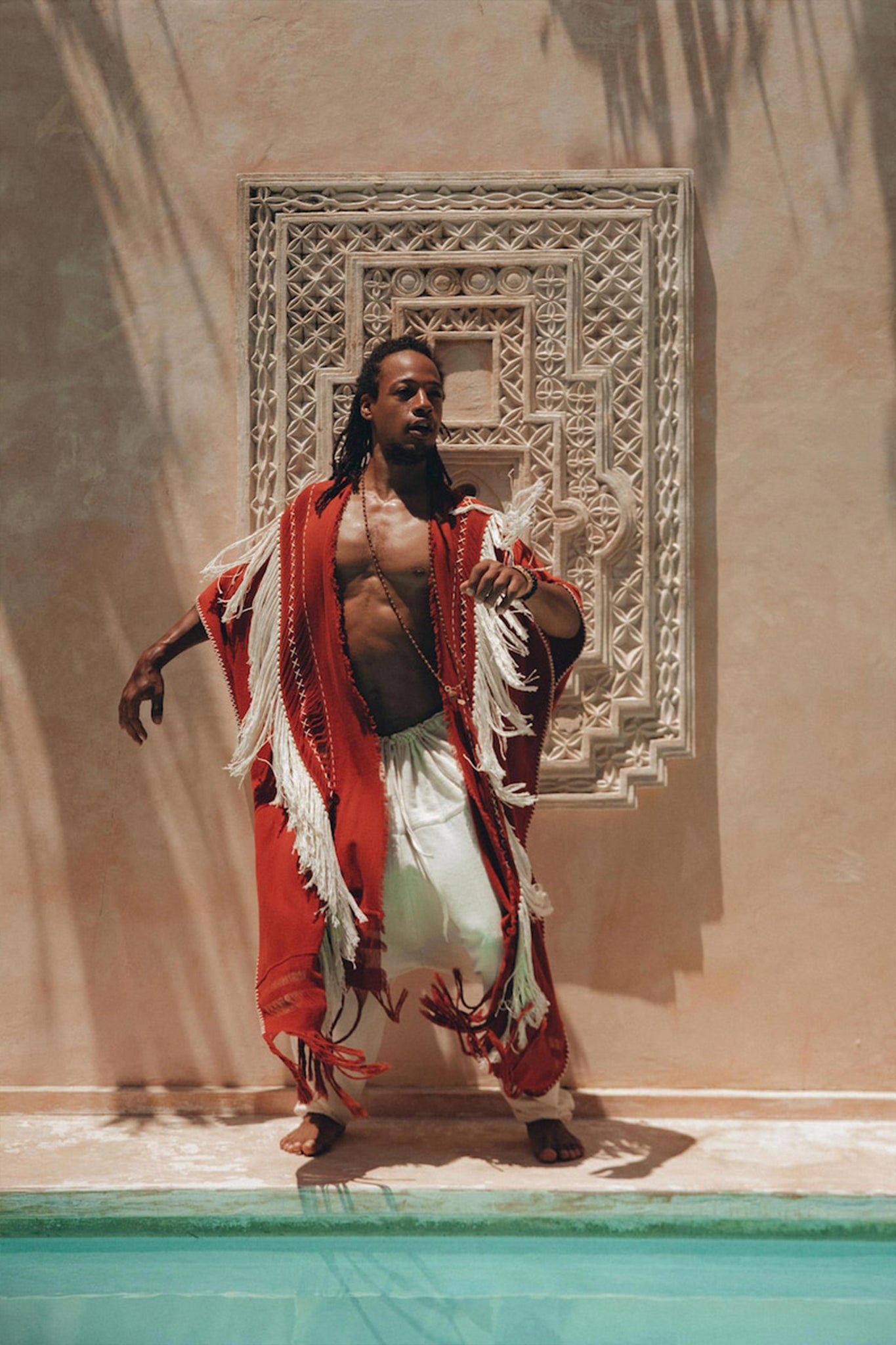 Wrapped in an AYA Sacred Wear Unisex Handwoven Cotton Wine Red Poncho and white pants, a person poses dramatically by a turquoise pool. Behind them, intricate white wall art creates a stunning backdrop as shadows of palm leaves enhance the shamanic tropical ambiance of the boho scene.