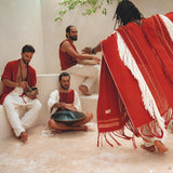 Four individuals adorned in red and white attire are assembled in a well-lit indoor area. One of them plays a hang drum, while another stands with their back to the camera, wearing an AYA Sacred Wear Unisex Handwoven Cotton Wine Red Poncho. They seem to be enjoying a laid-back musical gathering.