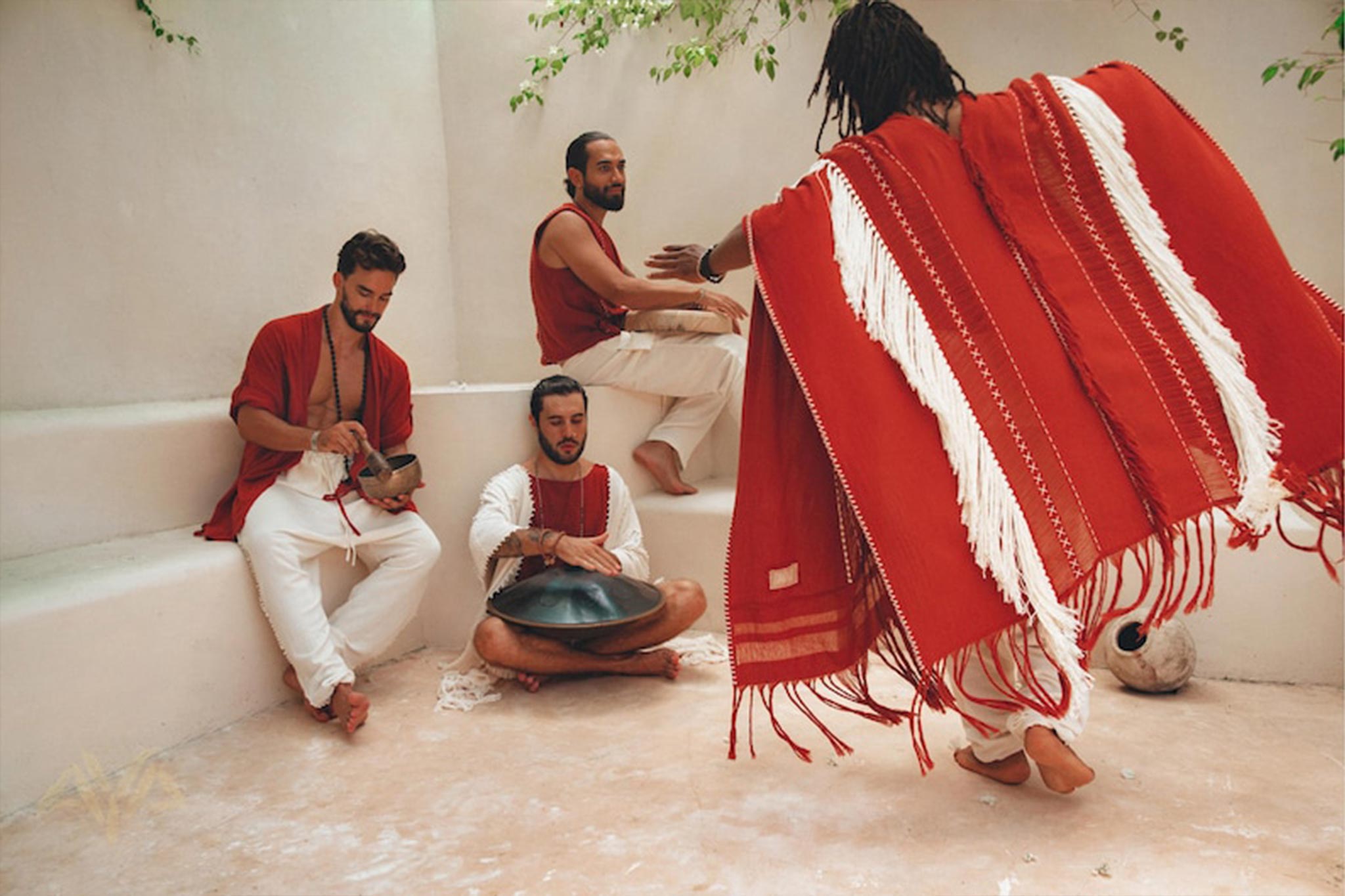 Four individuals adorned in red and white attire are assembled in a well-lit indoor area. One of them plays a hang drum, while another stands with their back to the camera, wearing an AYA Sacred Wear Unisex Handwoven Cotton Wine Red Poncho. They seem to be enjoying a laid-back musical gathering.