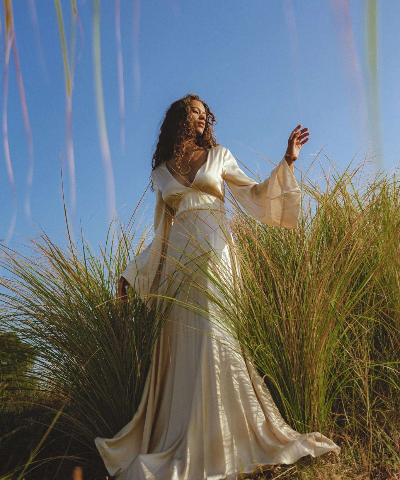 A woman in a Vanilla Cream Sundari Silk Gown by AYA Sacred Wear stands amid tall grass under a clear sky, extending her arm gracefully and gazing ahead, creating an elegant and ethereal scene.