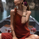 A woman in an AYA Sacred Wear Red Aurora Greek Goddess Dress, adorned with gold jewelry, sits cross-legged on a boat. She holds a purple flower to her ear and gazes to the side. The background features a serene water scene with cliffs and greenery, creating a tranquil and picturesque setting.