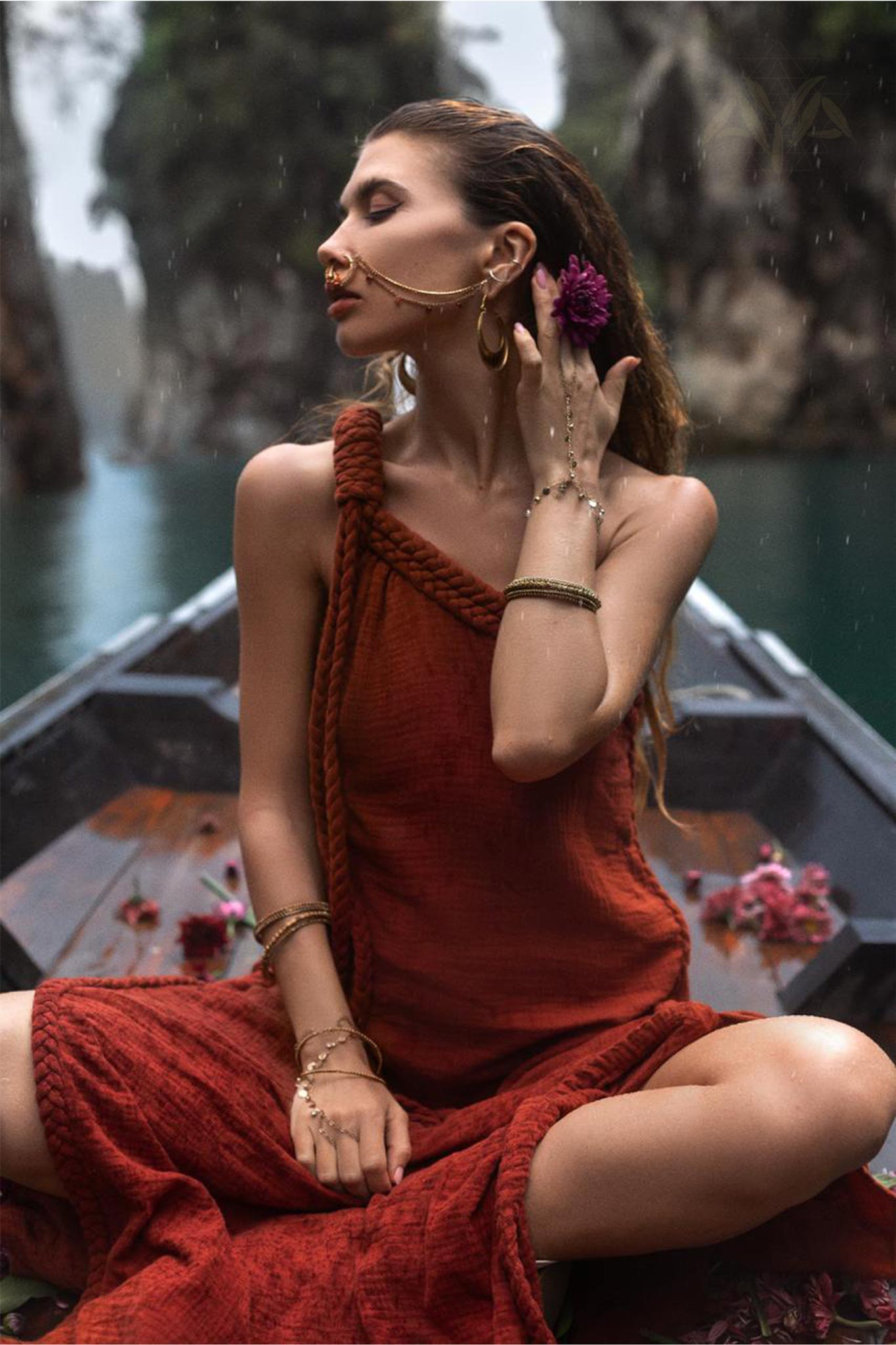 A woman in an AYA Sacred Wear Red Aurora Greek Goddess Dress, adorned with gold jewelry, sits cross-legged on a boat. She holds a purple flower to her ear and gazes to the side. The background features a serene water scene with cliffs and greenery, creating a tranquil and picturesque setting.