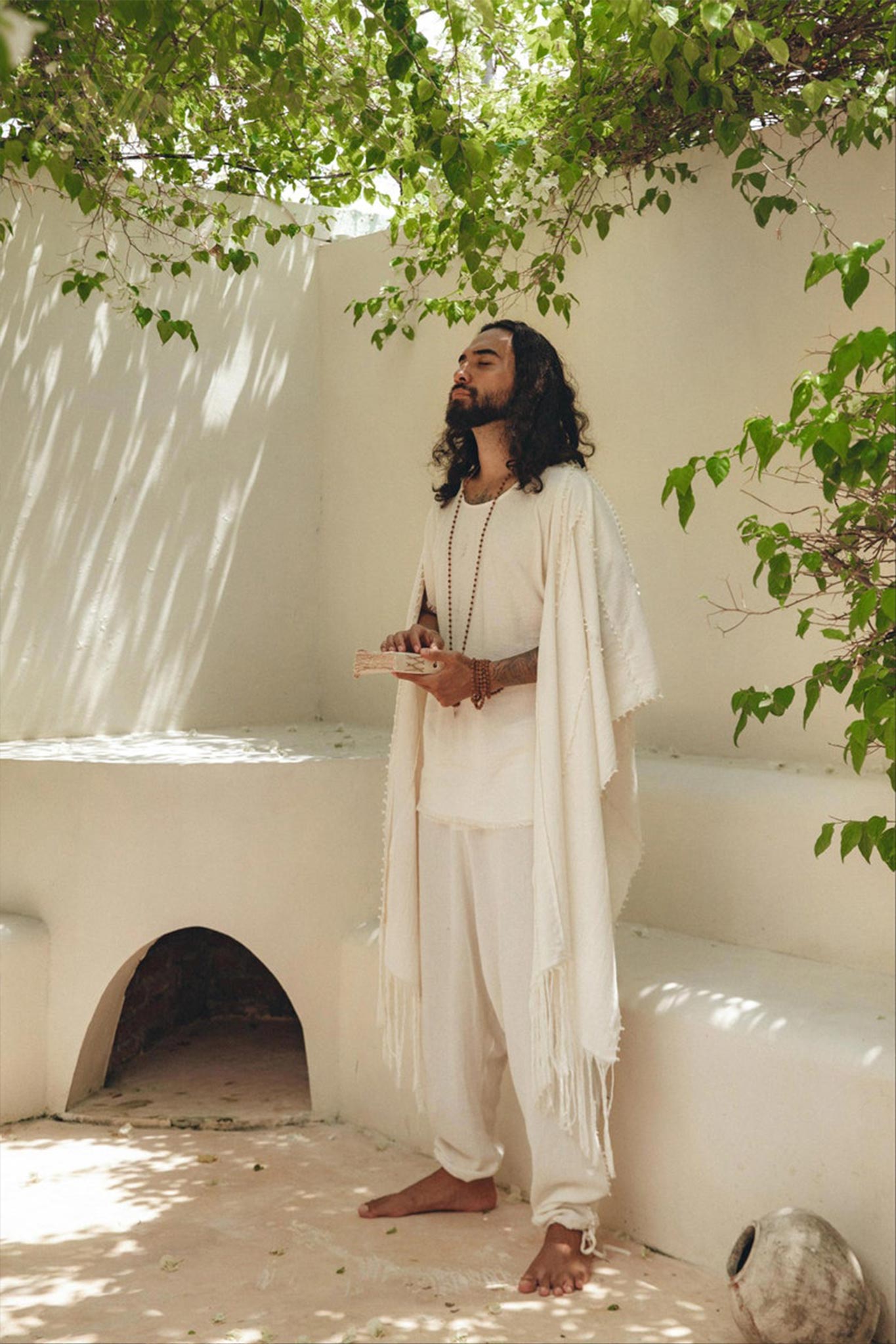 A person with long hair stands barefoot in a serene, sunlit courtyard, surrounded by leafy green branches. They are wearing an AYA Sacred Wear White Raw Cotton Minimalist Poncho for Men and holding a book, exuding a peaceful, contemplative presence.