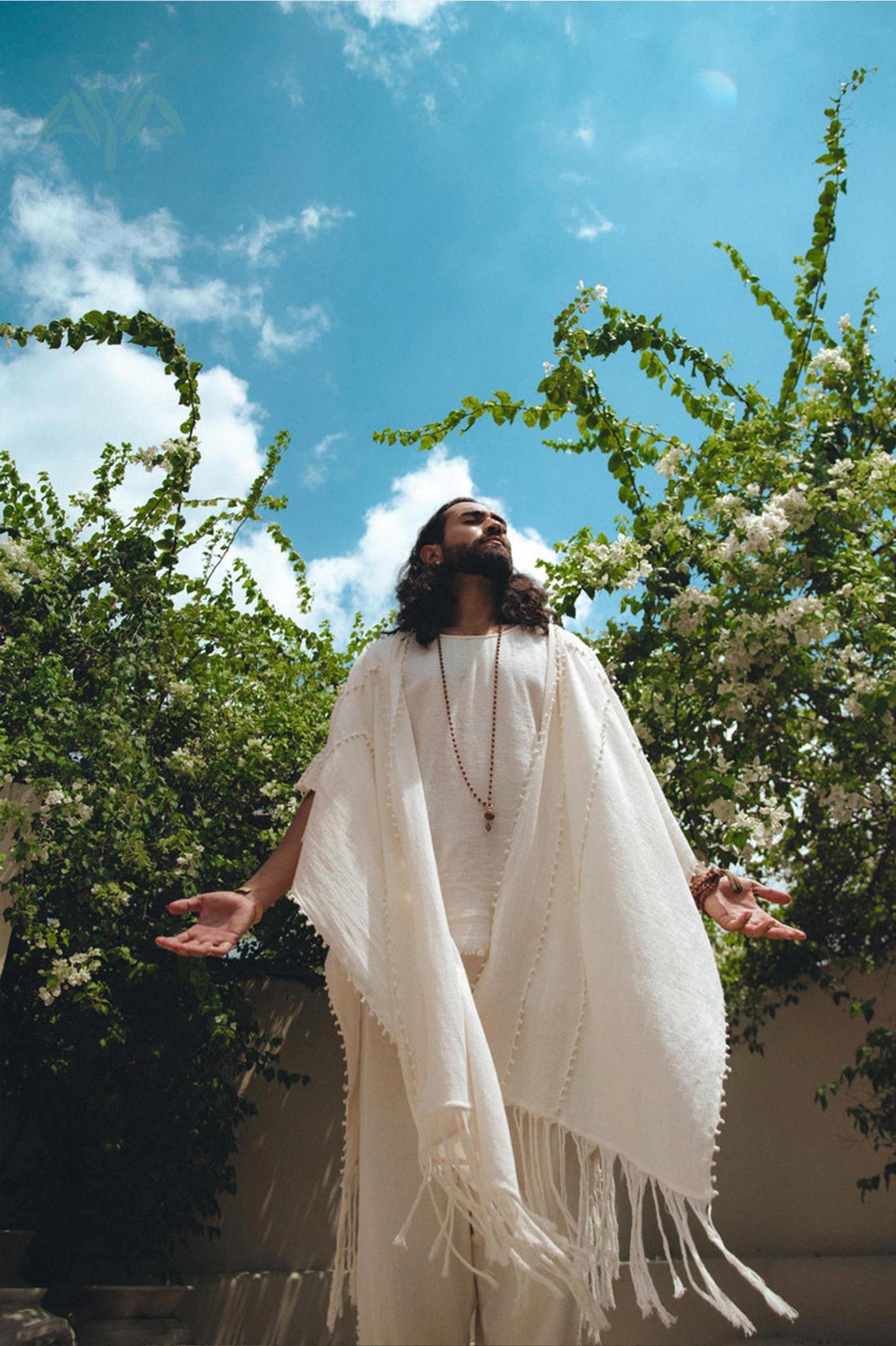 A figure with long hair stands outside, draped in a White Raw Cotton Minimalist Poncho for Men by AYA Sacred Wear. With arms open, they gaze at the bright blue sky. Green foliage and white flowers envelop them, enhancing the tranquil and serene setting. The botanically dyed handwoven cotton of the poncho perfectly harmonizes with the surrounding natural beauty.