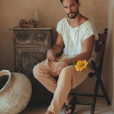 In a rustic room, a bearded man is seated on a chair, wearing the White Raw Edge Thick Cotton Top Tee from AYA Sacred Wear, paired with beige pants and sandals. A botanically dyed sunflower rests against his leg, complementing the serene atmosphere created by a wooden cabinet and large ceramic pot in the background.