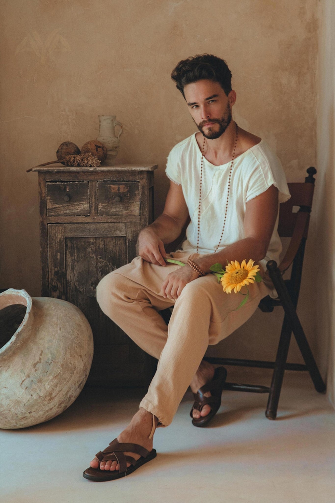 In a rustic room, a bearded man is seated on a chair, wearing the White Raw Edge Thick Cotton Top Tee from AYA Sacred Wear, paired with beige pants and sandals. A botanically dyed sunflower rests against his leg, complementing the serene atmosphere created by a wooden cabinet and large ceramic pot in the background.