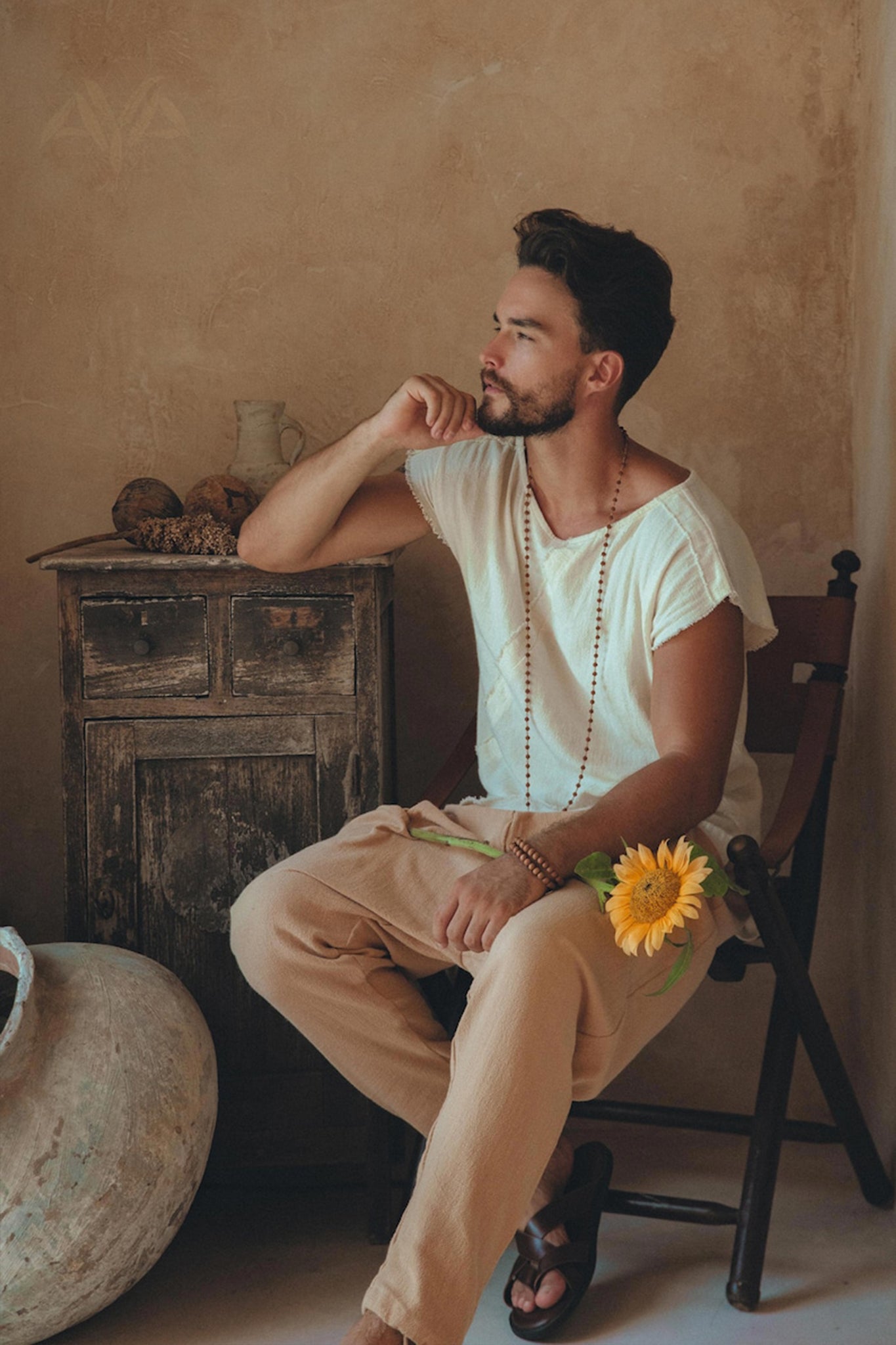 A man with a beard sits thoughtfully in a chair, wearing the White Raw Edge Thick Cotton Top Tee for Men by AYA Sacred Wear and beige pants. A sunflower peeks from his pocket and beads adorn his neck and wrist. The T-shirt, made from handwoven organic cotton, is botanically dyed. Behind him is a rustic wooden cabinet against a neutral-colored wall.