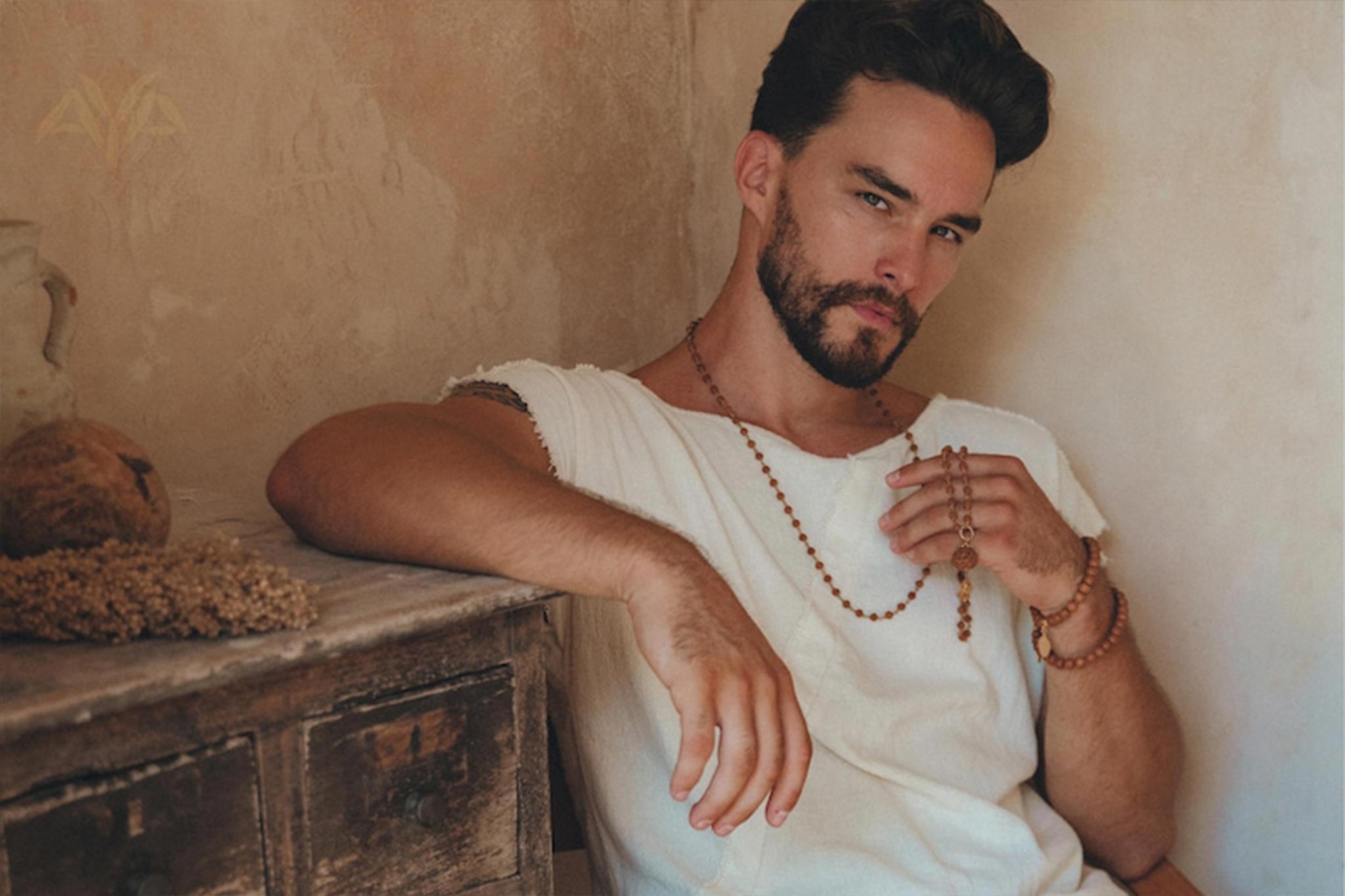 A man with a beard and wavy hair leans against a beige wall, wearing an AYA Sacred Wear White Raw Edge Thick Cotton Top Tee. He holds wooden prayer beads, while a rustic wooden chest adorned with natural decor sits beside him, enhancing the calm and earthy ambiance.