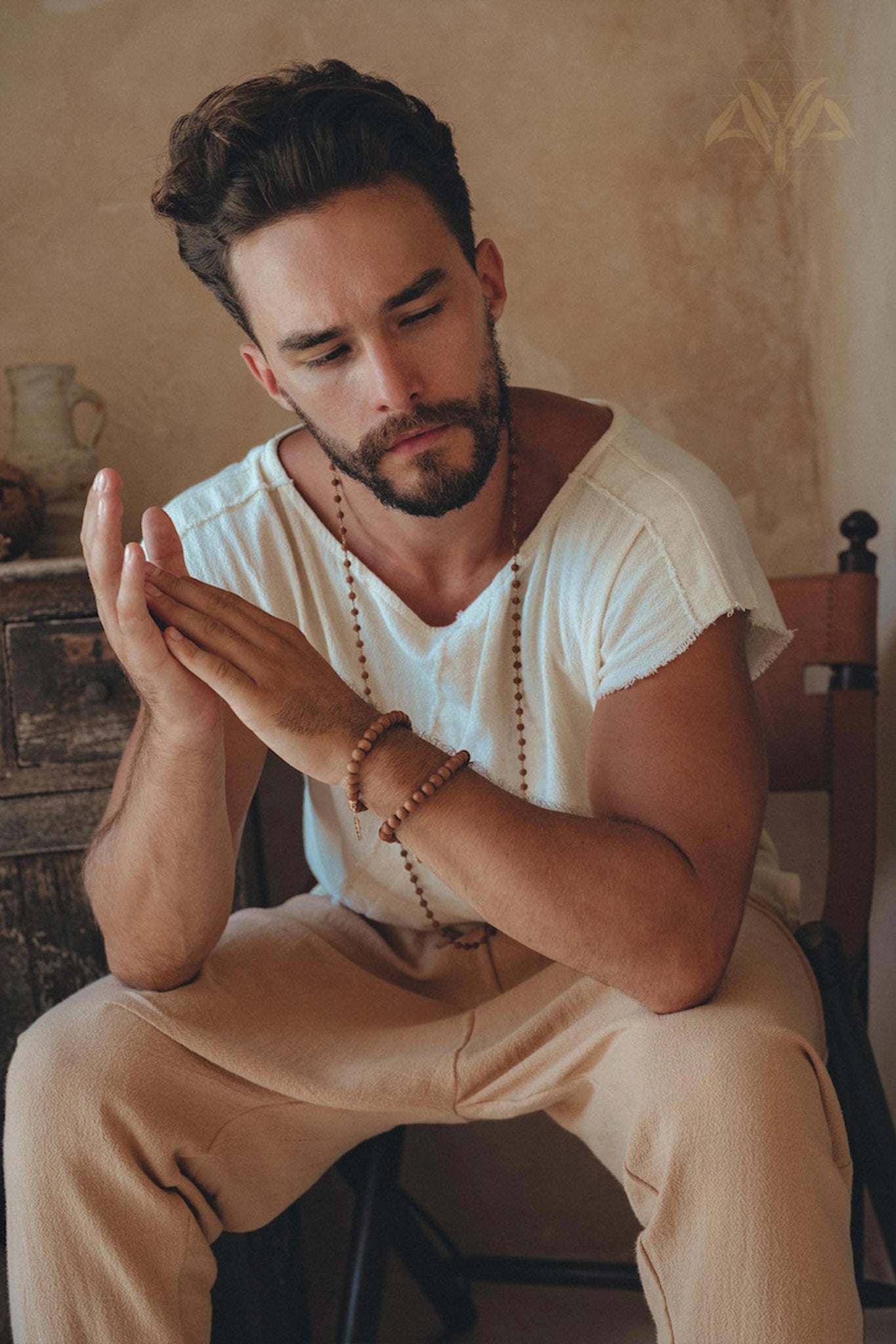 A man with a beard sits on a chair, dressed in a White Raw Edge Thick Cotton Top Tee by AYA Sacred Wear and cream-colored pants. His focused expression is accentuated by his hands clasped together, while a wooden necklace adorns his neck against the rustic backdrop of a wooden cabinet and a clay jug.