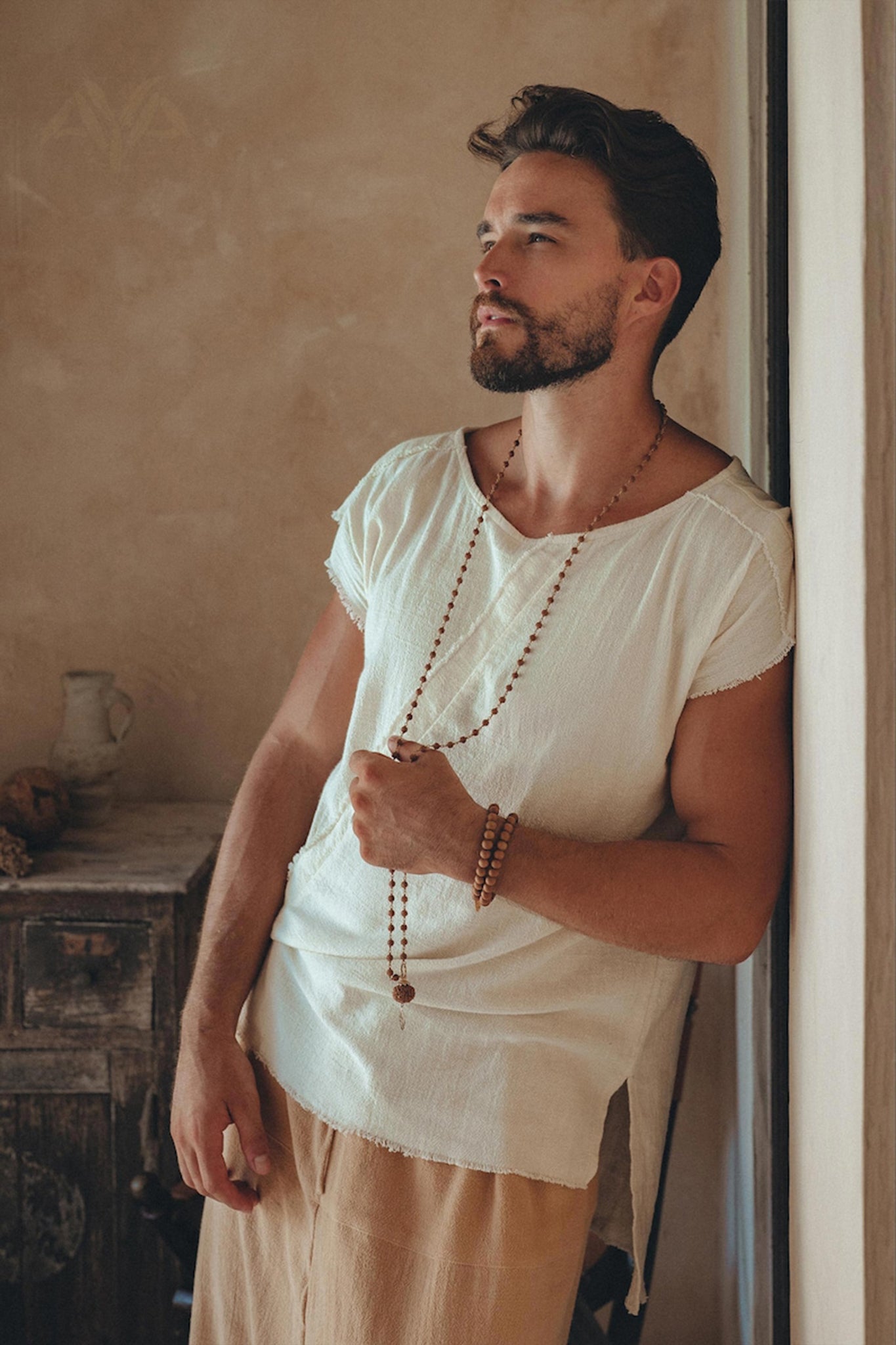 A man with a beard is wearing an AYA Sacred Wear White Raw Edge Thick Cotton Top Tee and beige pants, leaning against a rustic wall. He accessorizes with a beaded necklace and bracelet, displaying a contemplative expression. The background features earthy tones and a small wooden cabinet.