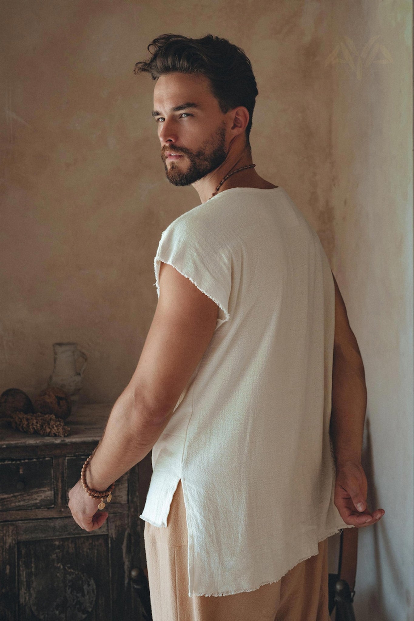 A man with a beard is indoors wearing a White Raw Edge Thick Cotton Top Tee from AYA Sacred Wear, and he is glancing over his shoulder. The background includes a rustic, textured wall and a wooden table adorned with decorative items.