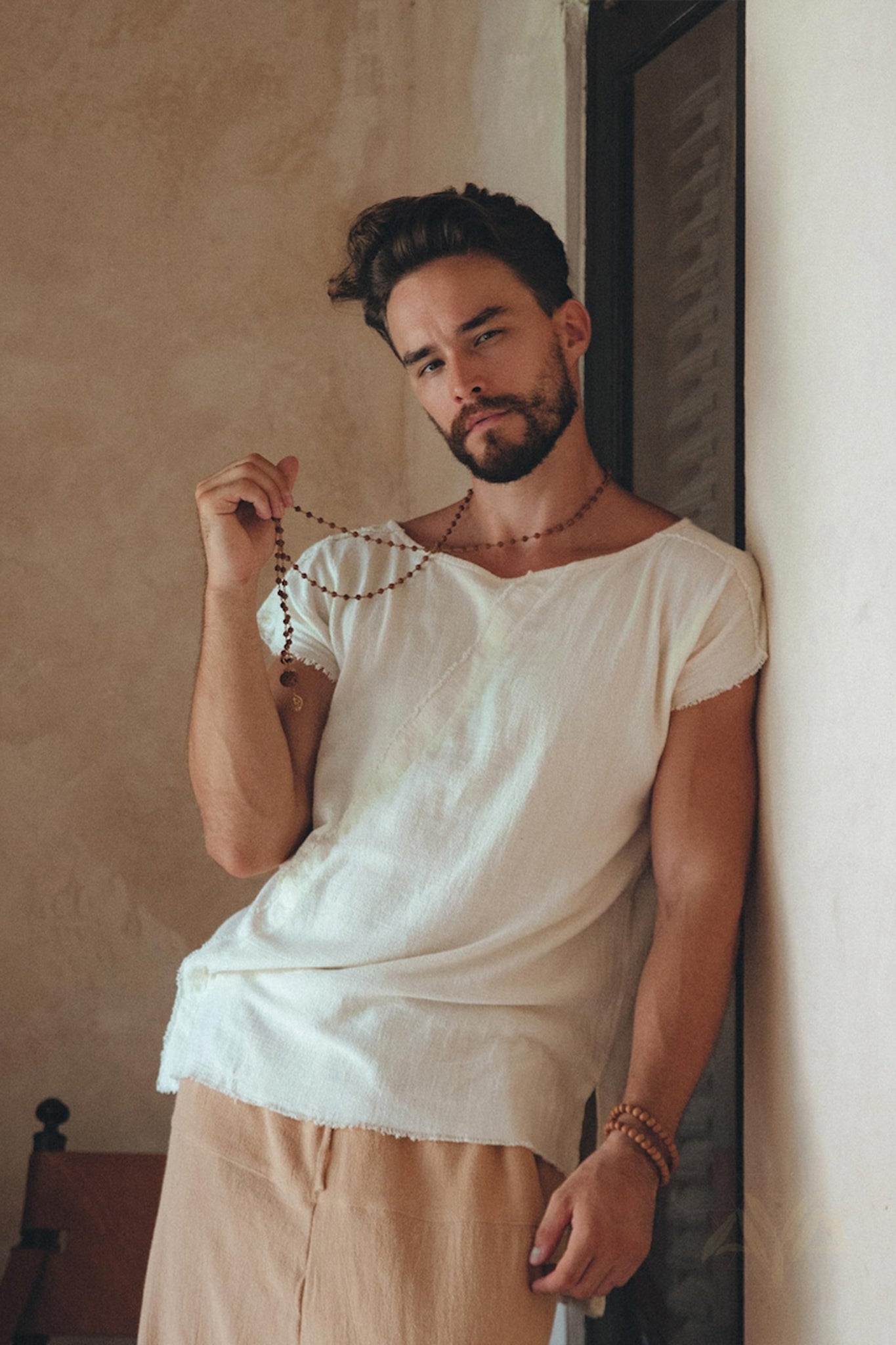 A bearded man in a relaxed, earth-toned setting leans against a wall, wearing an AYA Sacred Wear White Raw Edge Thick Cotton Top Tee for Men and beige pants while holding a string of beads. The minimalist ambiance enhances the tranquil atmosphere.