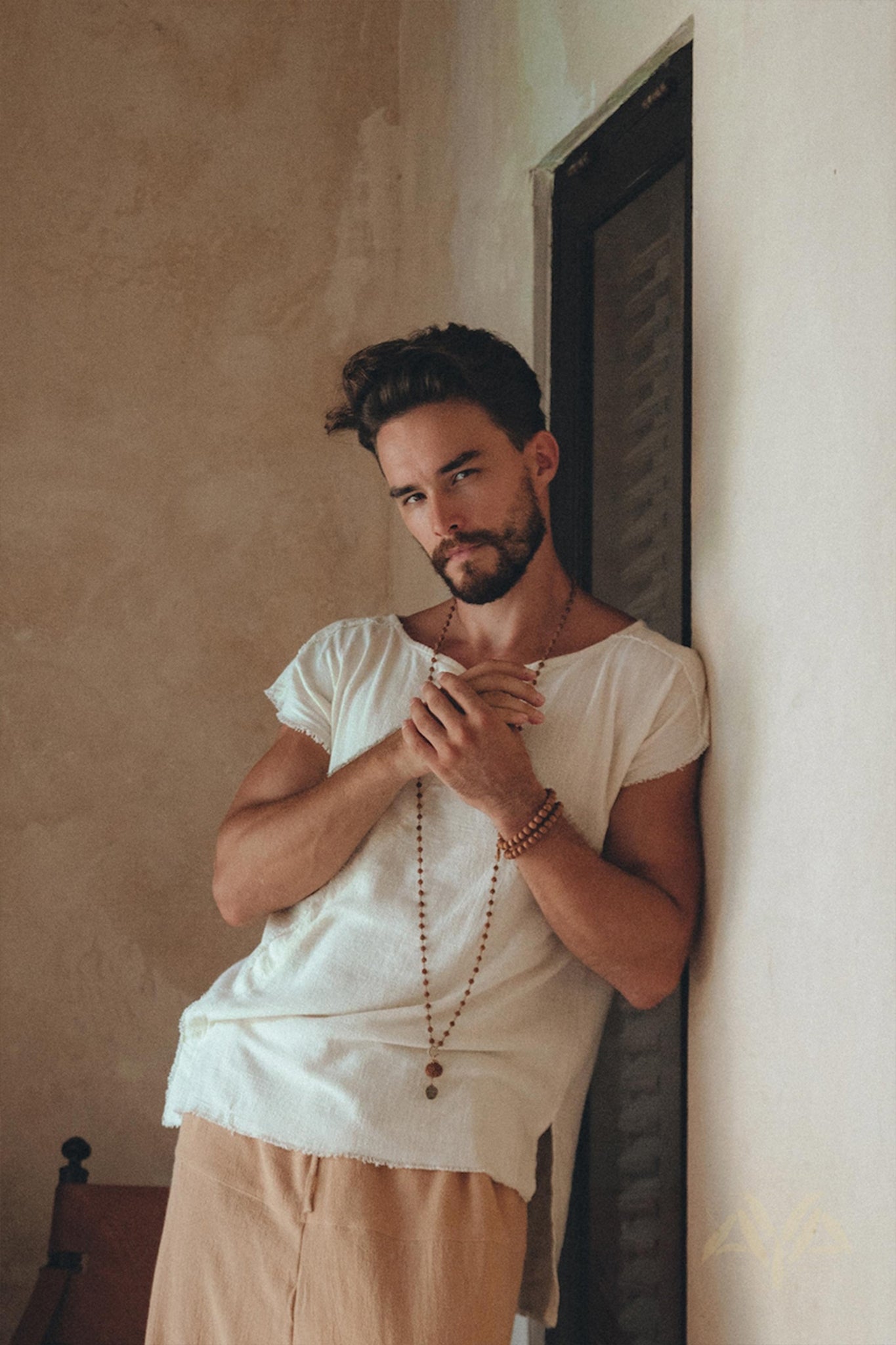 A man with a beard leans against a wall, wearing an AYA Sacred Wear White Raw Edge Thick Cotton Top Tee for Men paired with beige pants. He accessorizes with a long beaded necklace and bracelets, posing with hands together. The background is a textured, neutral-colored wall.