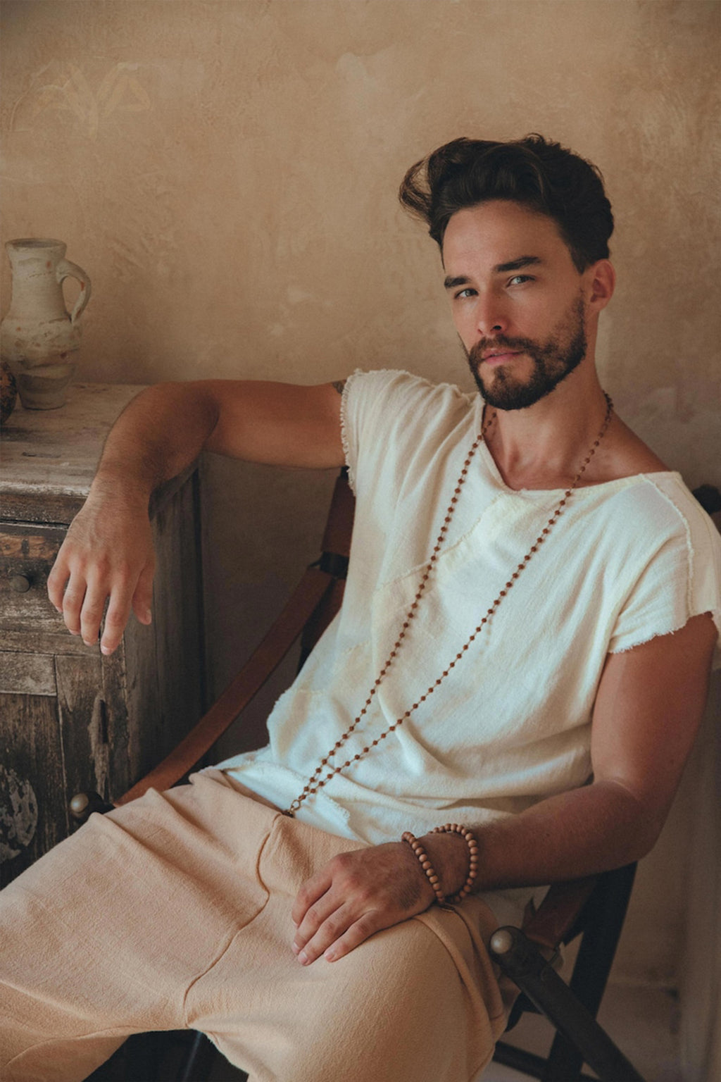 A bearded man with dark hair is sitting on a chair, wearing an AYA Sacred Wear White Raw Edge Thick Cotton Top Tee for Men and beige pants. He has a wooden bead necklace and bracelet, set against the backdrop of a rustic wall and a wooden chest adorned with pottery.