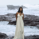 A woman in an AYA Sacred Wear Powder Blue Padma Dress stands on rugged ocean rocks with crashing waves and an overcast sky. Her long black hair cascades down her shoulders as she gazes ahead.