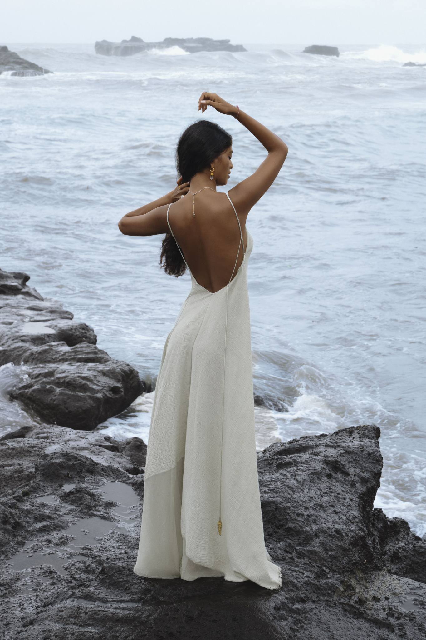 A woman wearing the Powder Blue Padma Dress by AYA Sacred Wear stands on rocky terrain by the ocean. Her long hair cascades down her back, and she raises one hand to touch her head, creating an elegant scene with waves in the background.