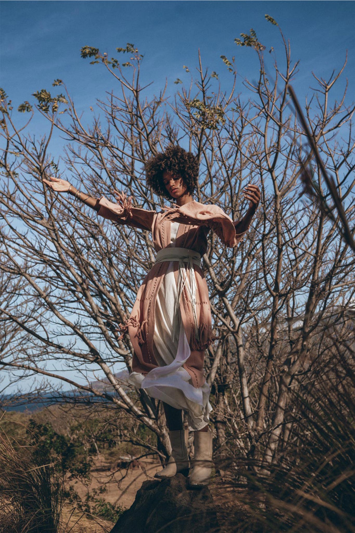 A person with curly hair stands gracefully on a rock, surrounded by leafless trees and tall grass. They wear a flowing beige and pink dress, cinched with the Woman’s White Leather Waist Belt from AYA Sacred Wear, paired with boots under the clear blue sky. The scene evokes a sense of tranquility and connection with nature.