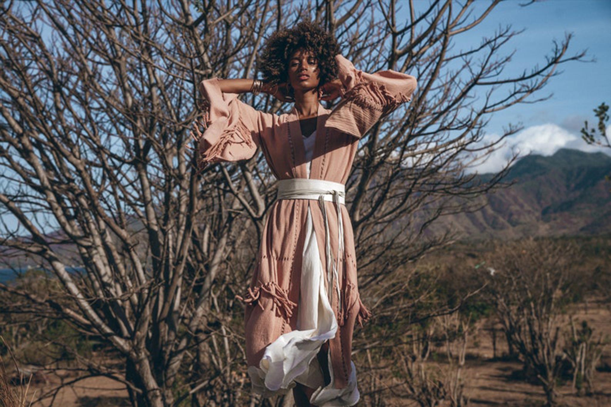 A person with curly hair stands outdoors, embodying a bohemian style in a long, flowing outfit paired with the Woman’s White Leather Waist Belt by AYA Sacred Wear. The handcrafted belt, made from pure organic genuine sheep leather, complements the natural setting of dry branches and a mountainous landscape under a clear sky.