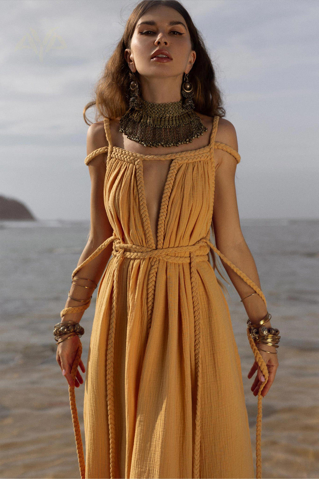 A woman stands on a beach wearing the NEW! Yellow Gold Greek Goddess Prom Boho Dress from AYA Sacred Wear. Her long brown hair complements the large, intricate metal necklace and multiple bracelets she wears. In the background, the sea and a headland create a tranquil backdrop for her enchanting attire.