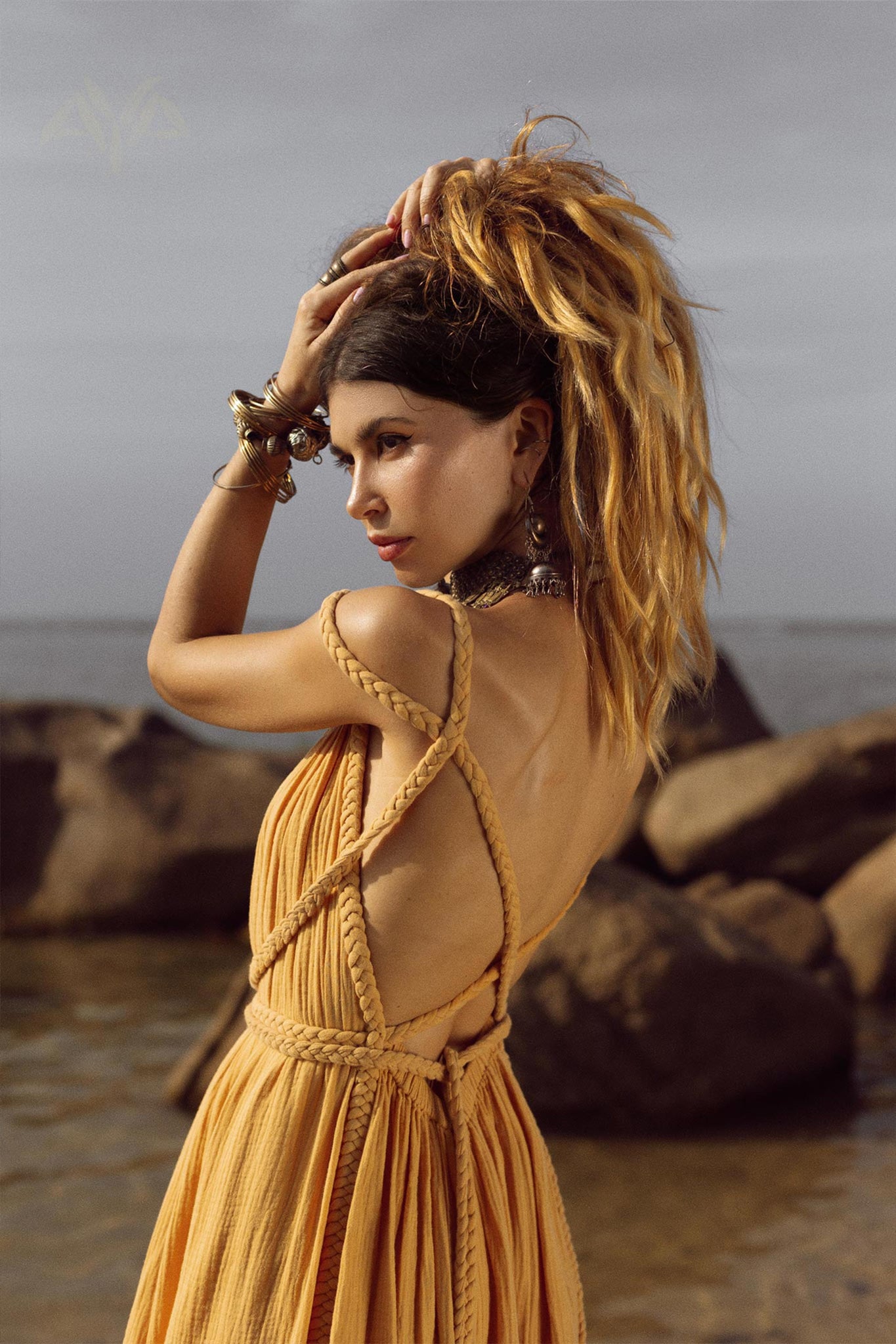 A woman with long, wavy hair tied up poses on a rocky beach, wearing the NEW! Yellow Gold Greek Goddess Prom Boho Dress by AYA Sacred Wear, featuring a backless design, organic cotton fabric, and rope details. She accessorizes with several bracelets while the overcast sky and sea create a dramatic background.