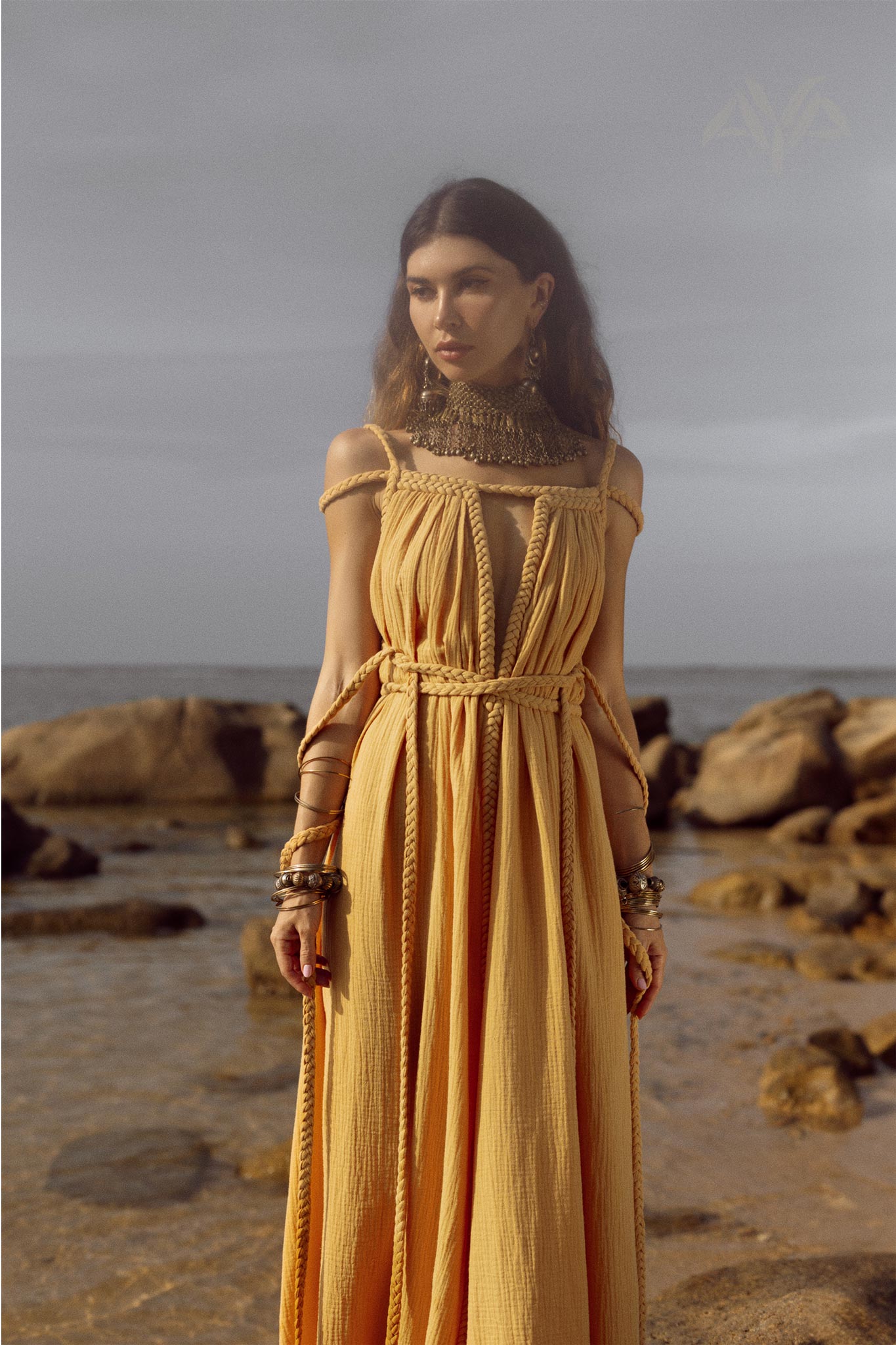 On a rocky beach, a woman is seen wearing the NEW! Yellow Gold Greek Goddess Prom Boho Dress by AYA Sacred Wear. The dress, with its intricate strap designs, evokes a boho wedding style. Her look is enhanced by jewelry, including bold necklaces and bracelets. Above her, the sky is overcast while the sea remains calm in the background.