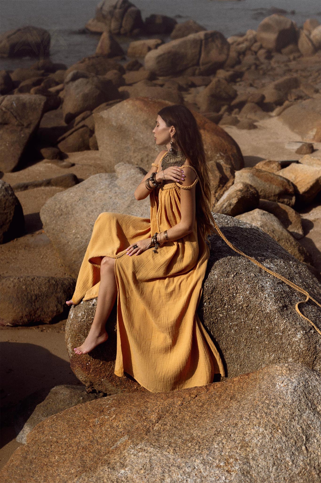 A woman in a flowing Greek Goddess Prom Boho Dress from AYA Sacred Wear sits on large rocks by the sea. She has long hair and wears layered jewelry, creating a natural, serene backdrop against the beach and rocky shoreline.