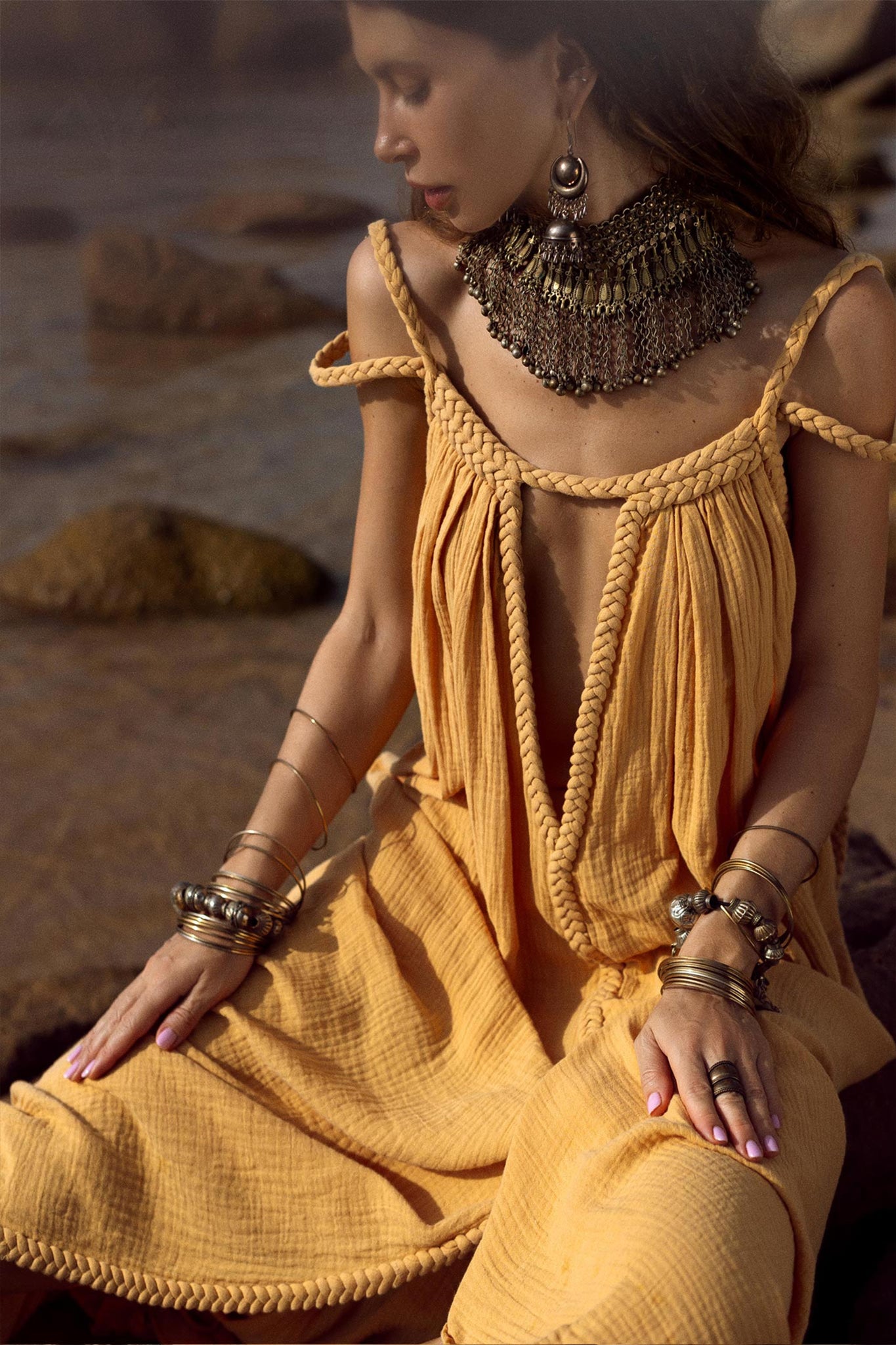 A woman in an AYA Sacred Wear NEW! Yellow Gold Greek Goddess Prom Boho Dress with braided straps and a large metal necklace sits on rocks by the water. Her head is slightly turned from the camera, bathed in warm, natural light. Multiple bracelets adorn her wrists, enhancing her boho-chic vibe.