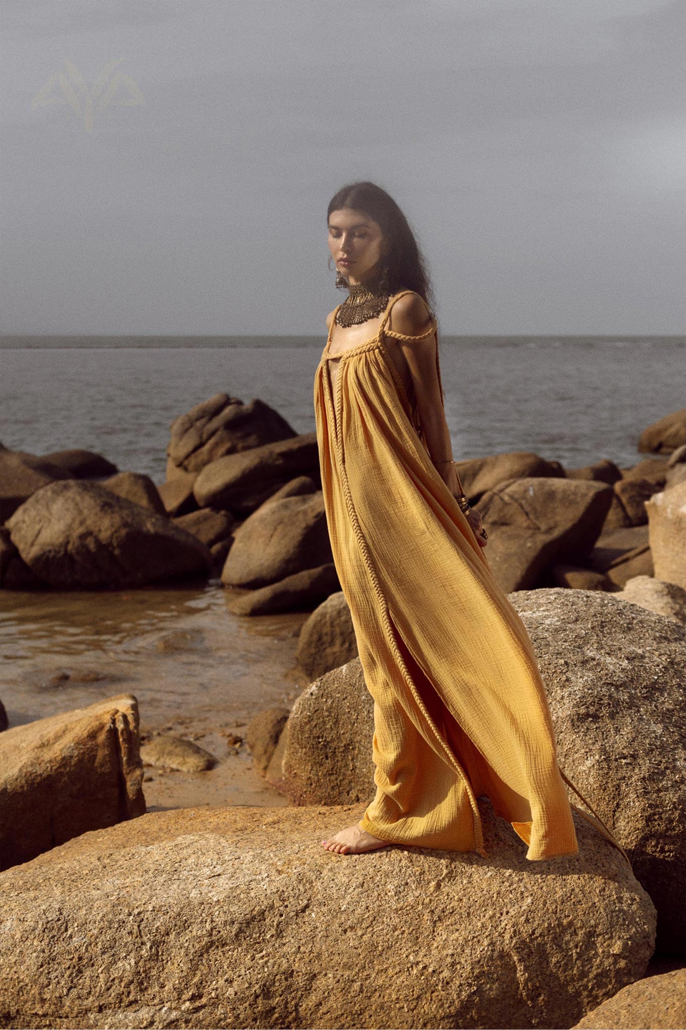 A person wearing the NEW! Yellow Gold Greek Goddess Prom Boho Dress from AYA Sacred Wear stands barefoot on large rocks by the sea. The overcast sky and calm water together create a serene and peaceful atmosphere.