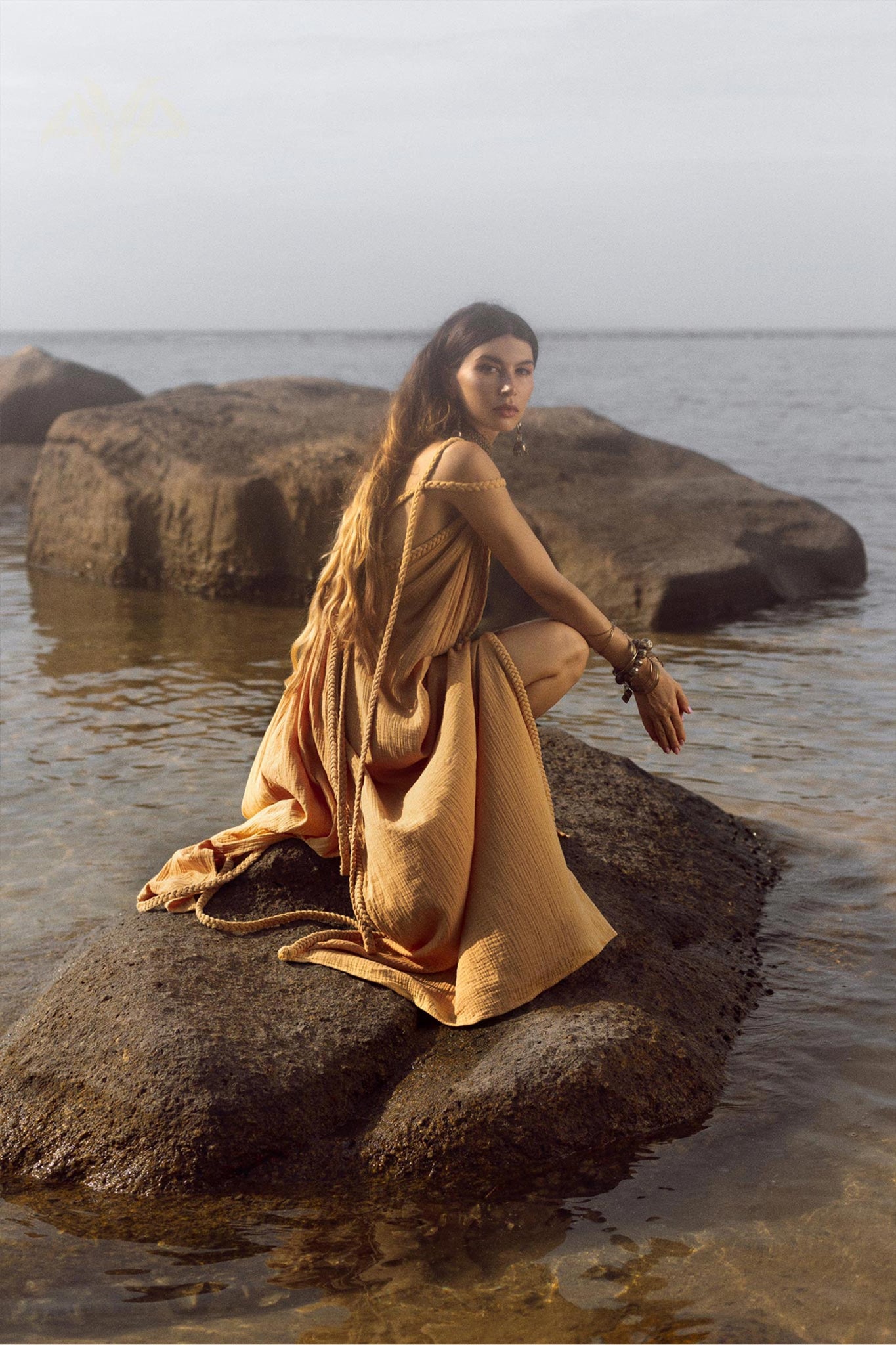 A woman in the NEW! Yellow Gold Greek Goddess Prom Boho Dress by AYA Sacred Wear crouches on a large rock by the sea. The calm water and additional rocks in the background enhance the serene, natural setting.