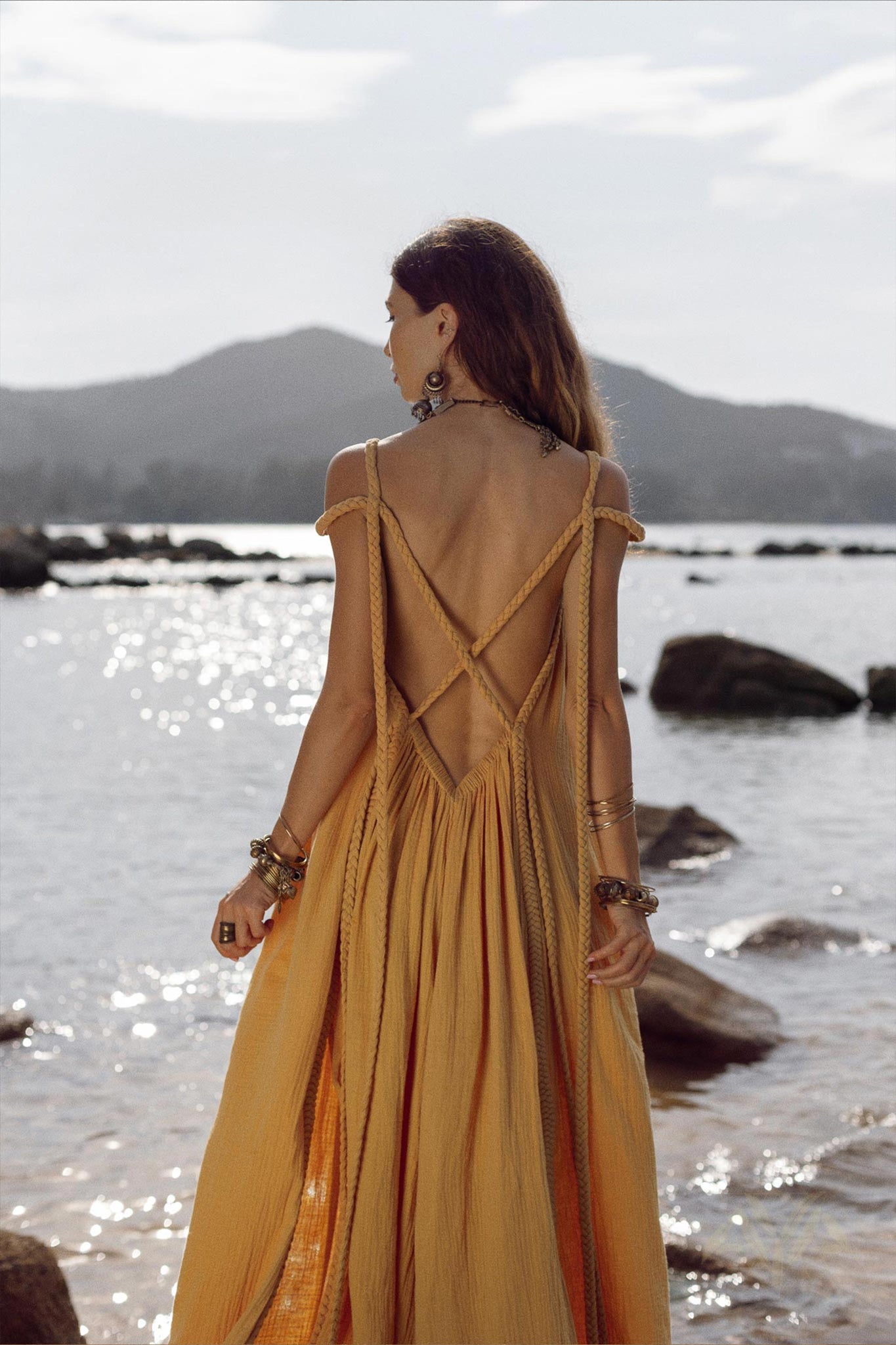 A woman wearing the NEW! Yellow Gold Greek Goddess Prom Boho Dress by AYA Sacred Wear stands beside a rocky shoreline, gazing out at the water. The dress features an intricate open-back design that captures the elegance of a boho wedding gown. Sunlight reflects off the water in the bright scene, with distant mountains visible in the background.