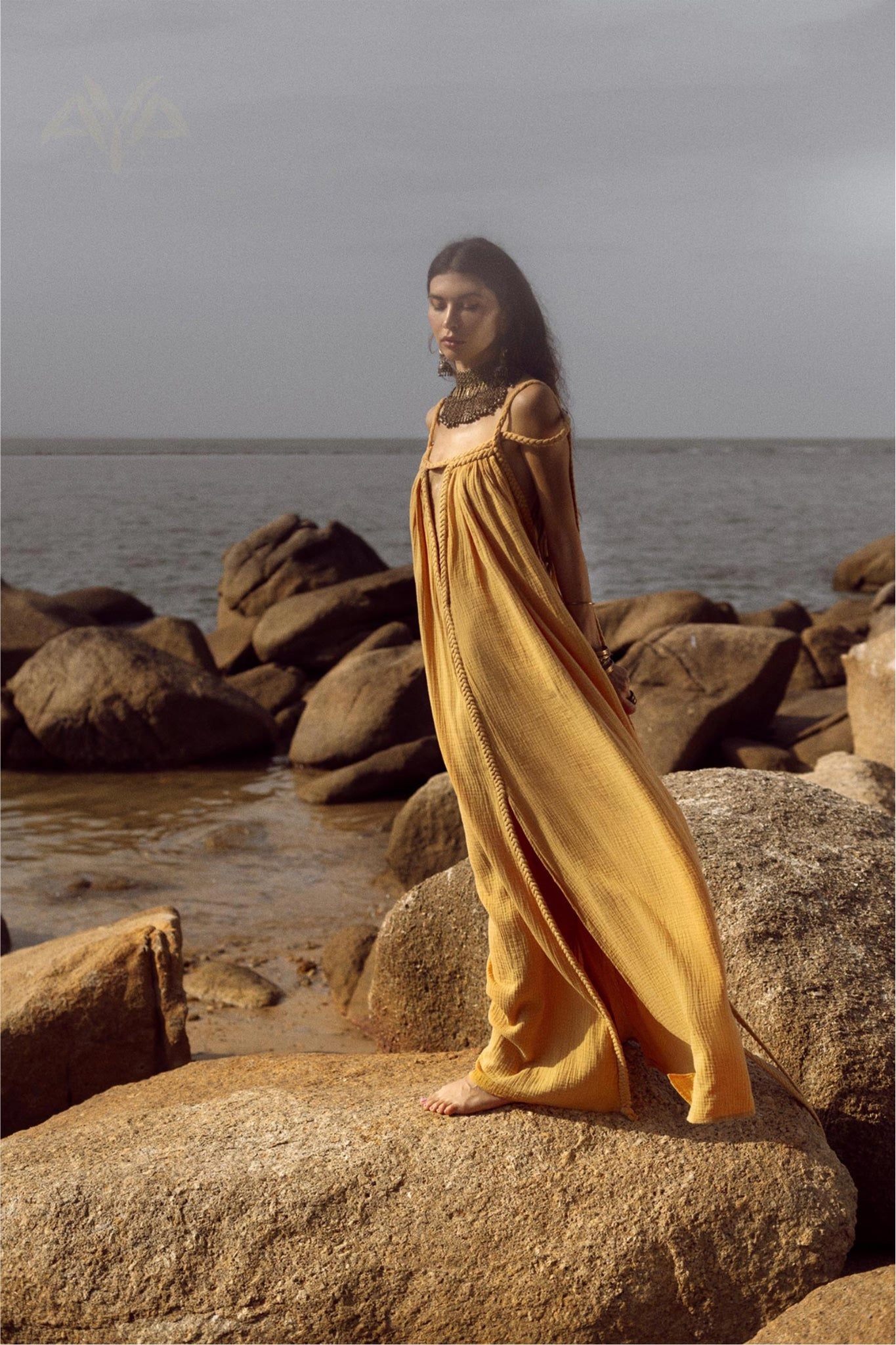 A woman in the Yellow Gold Greek Goddess Prom Boho Dress by AYA Sacred Wear stands barefoot on rocks by the sea. Her long dark hair flows as she gazes into the distance, with an overcast sky above and a calm ocean forming a serene backdrop. Perfect for special occasions.