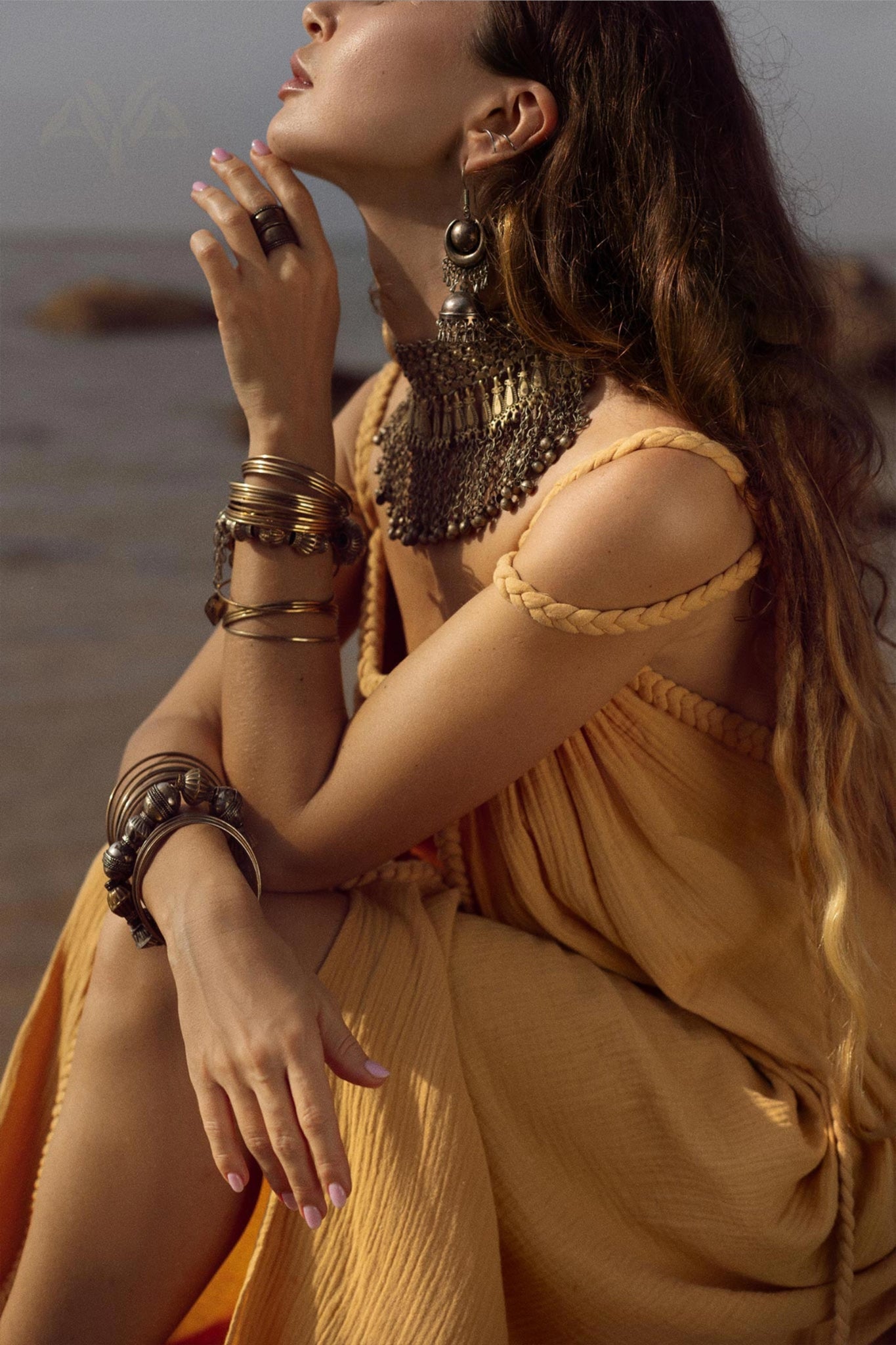 A woman in an AYA Sacred Wear Yellow Gold Greek Goddess Prom Boho Dress sits by the beach, adorned with intricate jewelry including a necklace, hoop earrings, and bracelets. Her long brown hair cascades down her back as she gazes upwards.