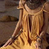 A woman in an AYA Sacred Wear Yellow Gold Greek Goddess Prom Boho Dress, with braided straps, sits on rocks. The lightweight cotton dress is ideal for special occasions. She wears a metallic necklace and bangles, her pink nails glowing in the sunlit backdrop.