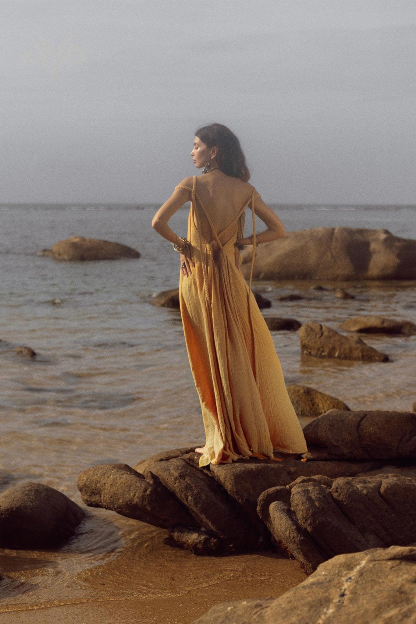 A woman in an AYA Sacred Wear Yellow Gold Greek Goddess Prom Boho Dress stands on coastal rocks, gazing at the ocean. The serene scene features gentle waves and a clear sky. Ideal for special occasions, the dress embodies nature's beauty and tranquility.